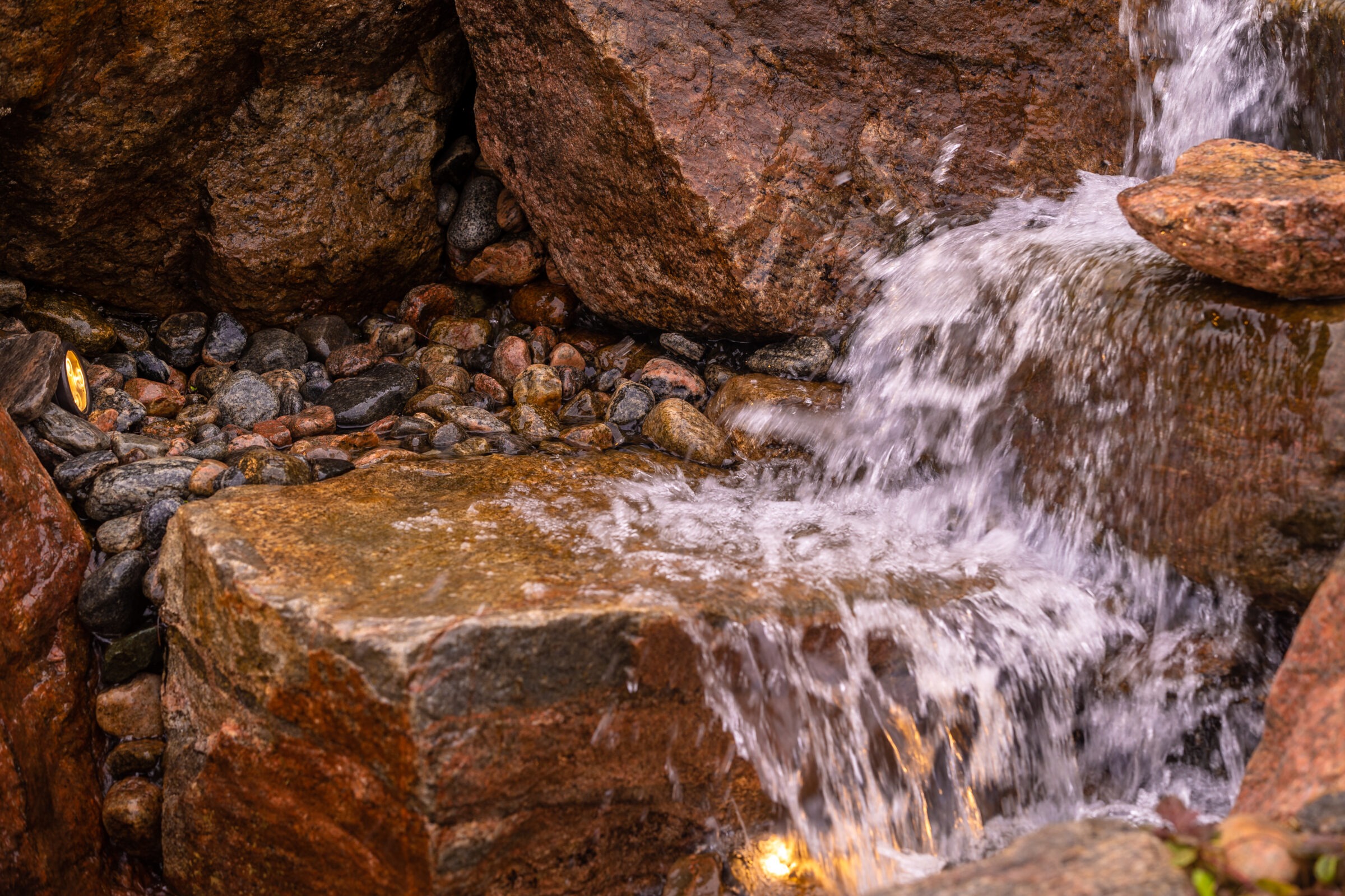 A cascading waterfall flows over natural rocks, surrounded by smooth pebbles, with warm lighting illuminating the serene, earthy scene.