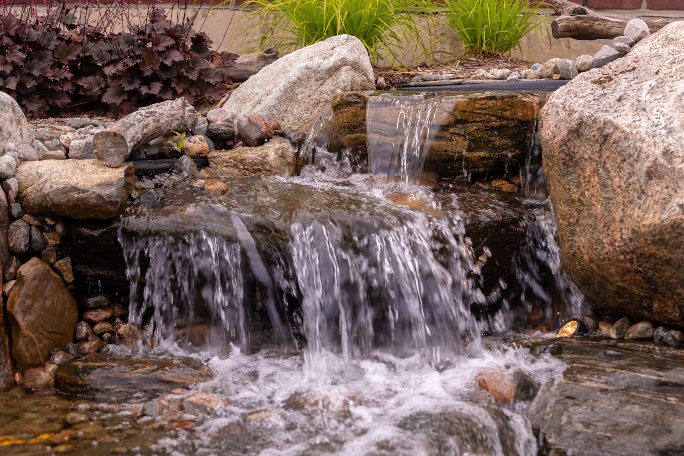 Small waterfall cascading over rocks, surrounded by greenery and stones, creating a tranquil natural scene without any visible buildings or people.