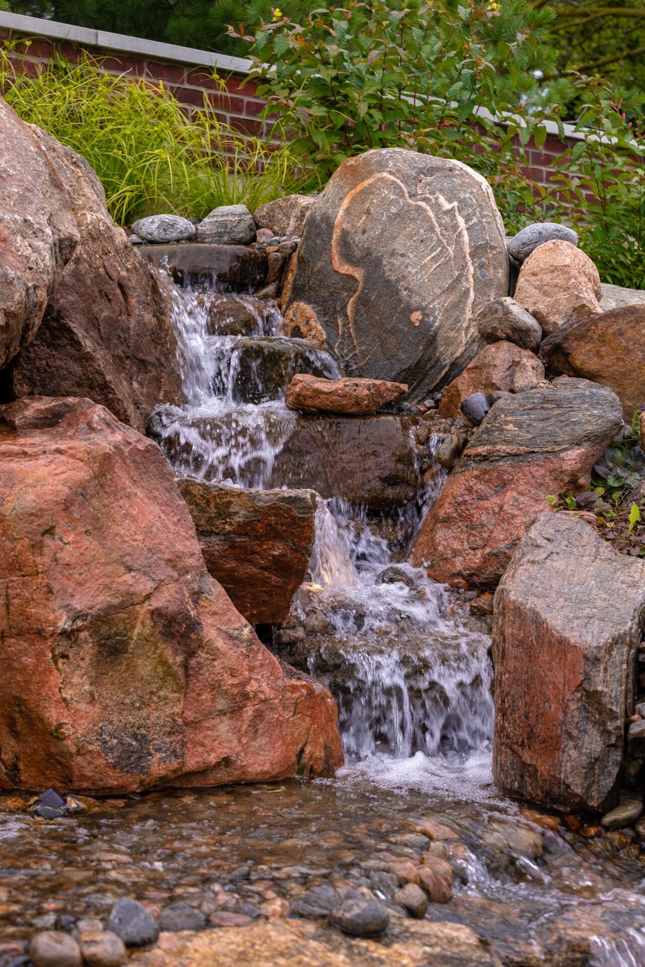A small waterfall cascades over large rocks, surrounded by green plants. A brick wall is visible in the background.