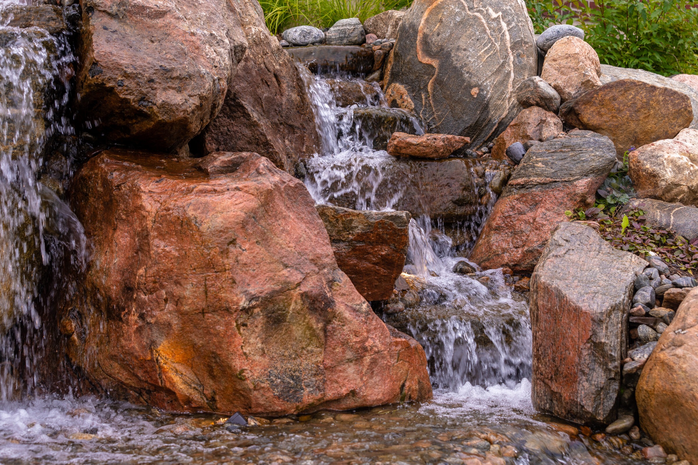 A small waterfall flows over large rocks, surrounded by greenery. Water cascades gently down, creating a peaceful, natural scene.