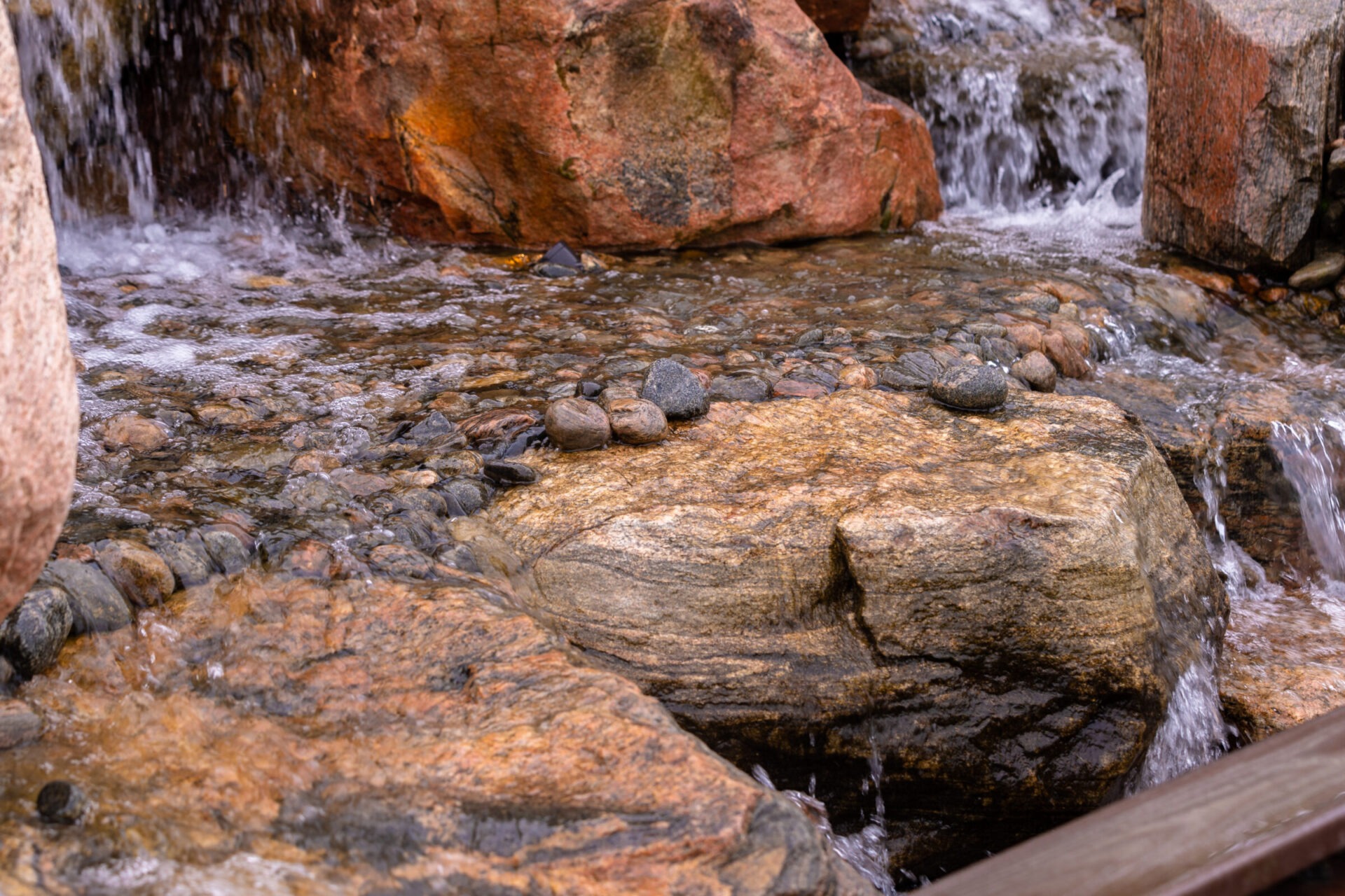 A small waterfall flows over rocks, creating a serene, natural scene with clear water cascading gently over smooth stones and textured boulders.