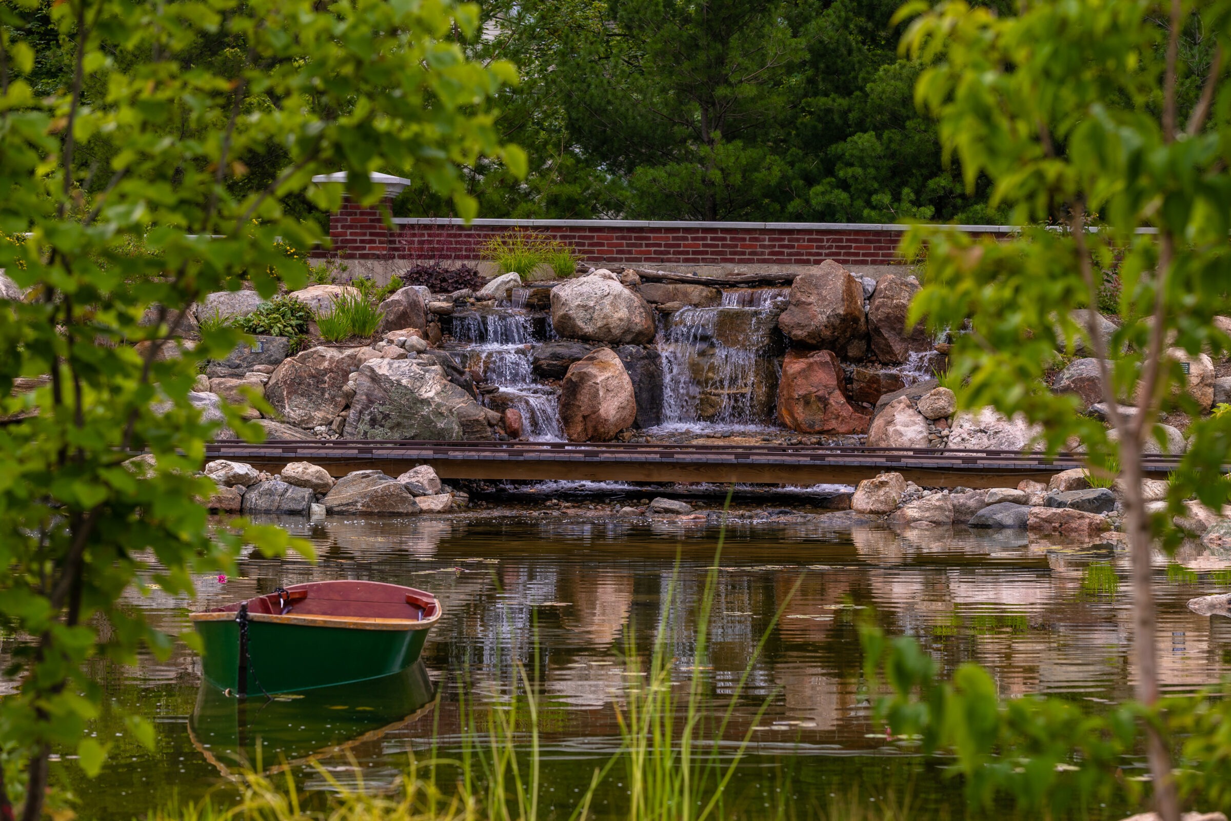 A serene garden scene with a small waterfall, lush greenery, a green boat on a pond, and a low brick wall backdrop.
