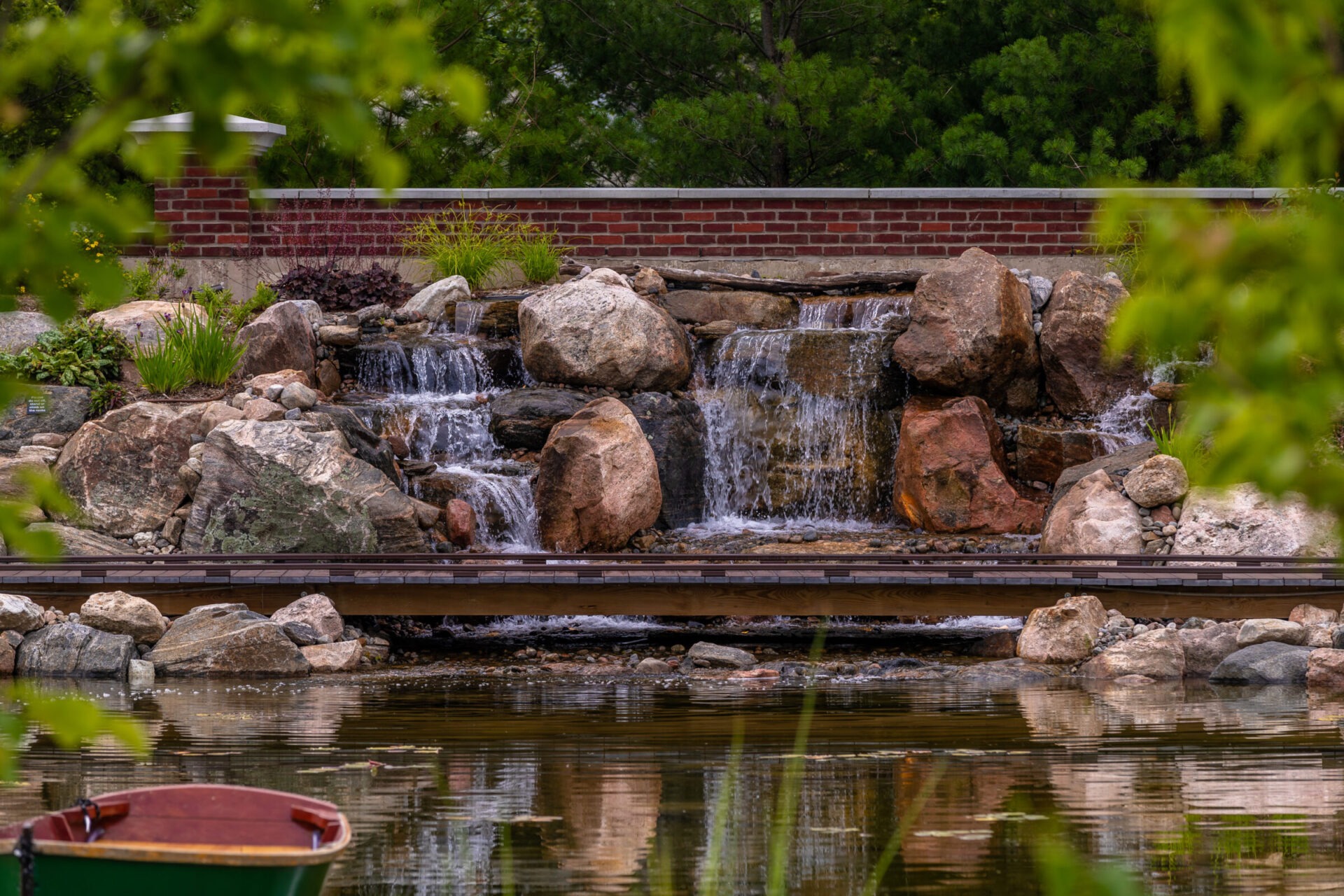 A serene garden scene shows a small waterfall cascading over rocks into a pond, surrounded by lush greenery and a wooden bridge.