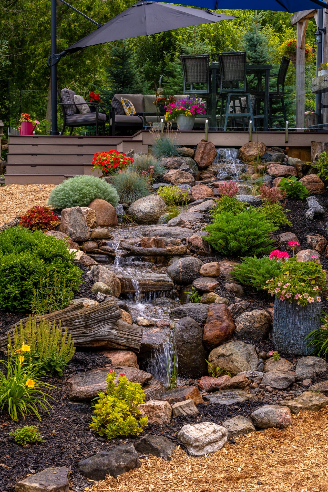 A tranquil backyard patio features lush greenery, a small waterfall, colorful flowers, and seating under umbrellas, creating a serene outdoor retreat.