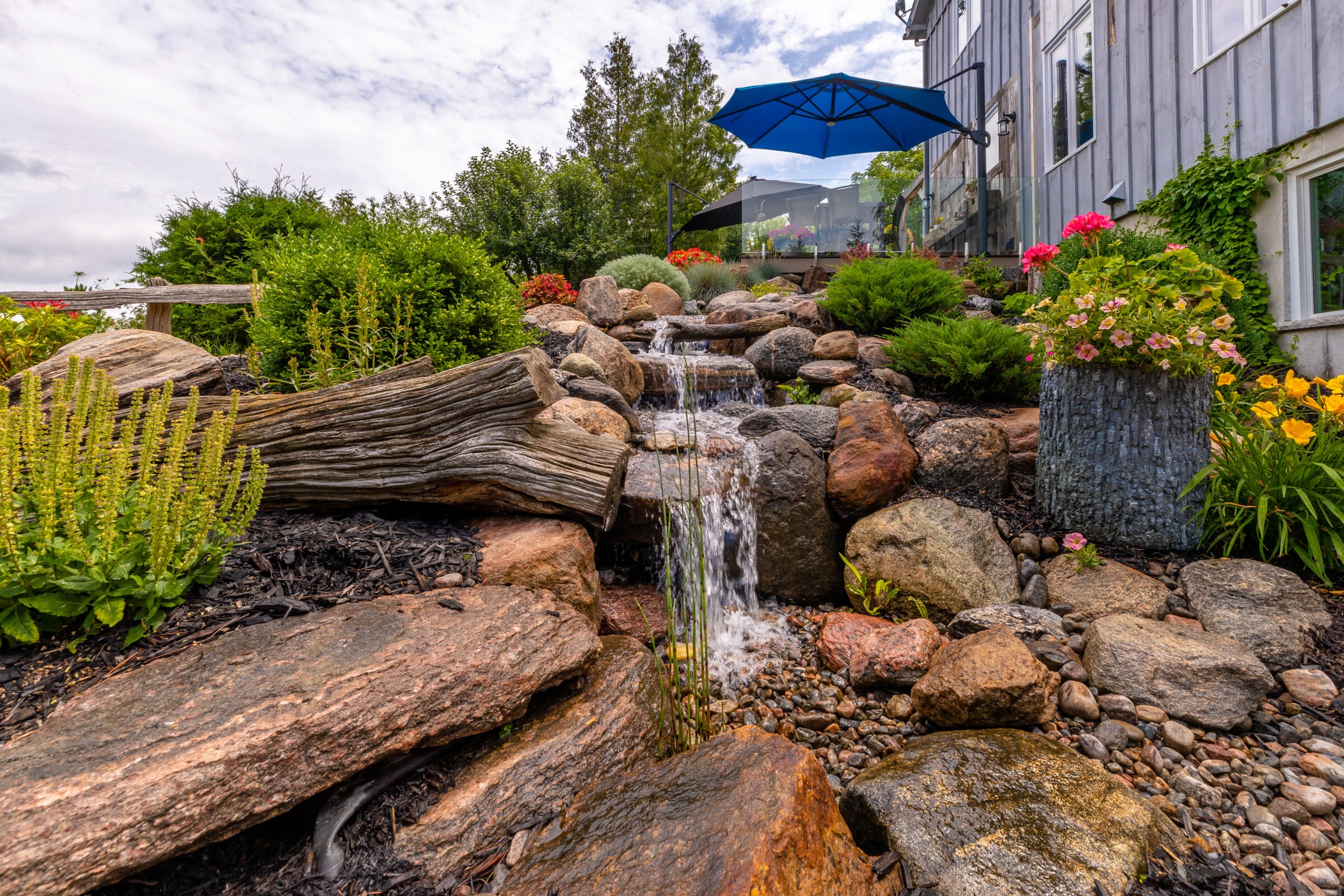 A cascading garden waterfall with vibrant flowers surrounds a stone path. Blue umbrella and house are visible in the background.