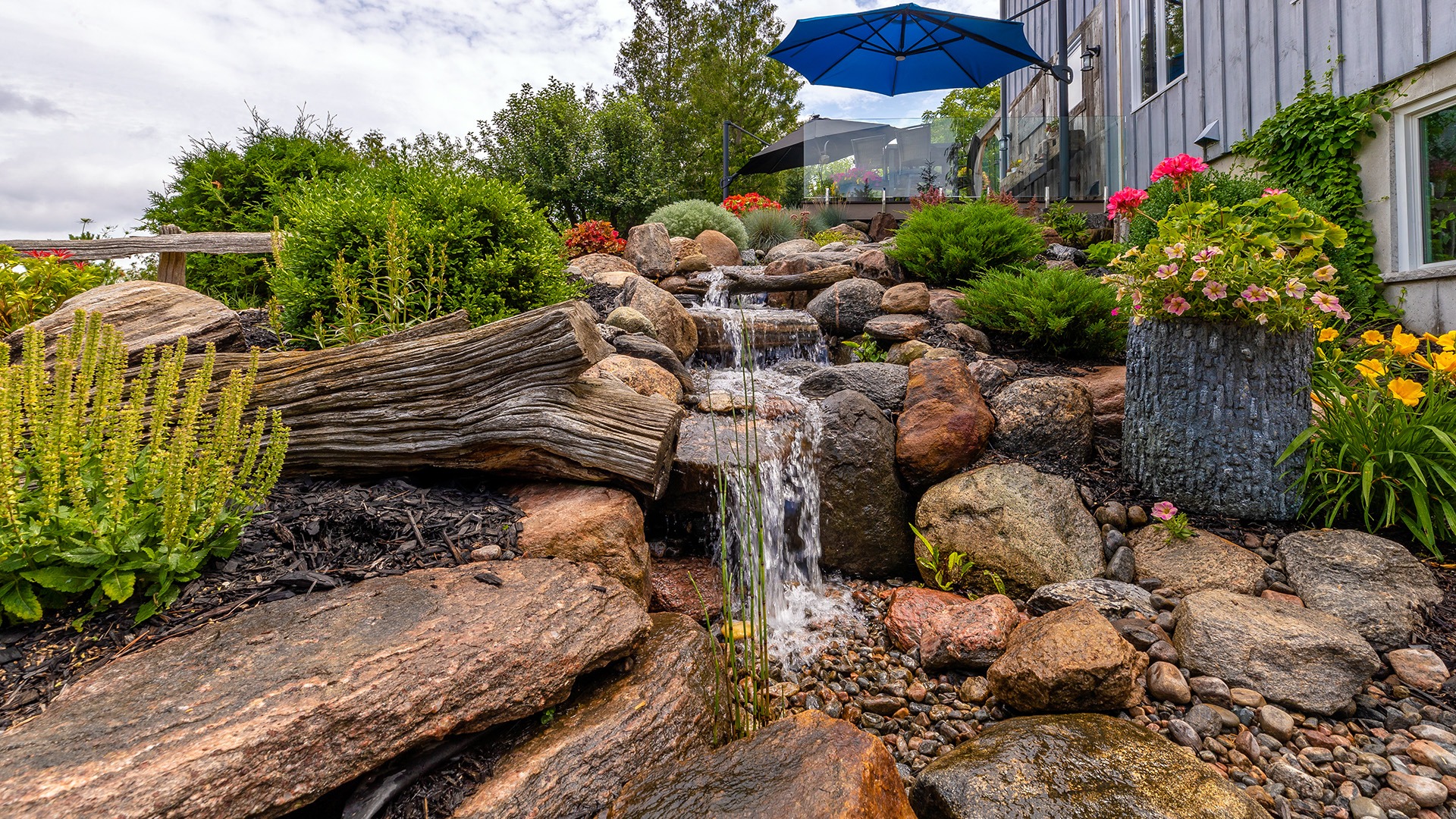 A small waterfall flows over rocks in a lush garden, surrounded by vibrant flowers and greenery, under a blue umbrella and cloudy sky.