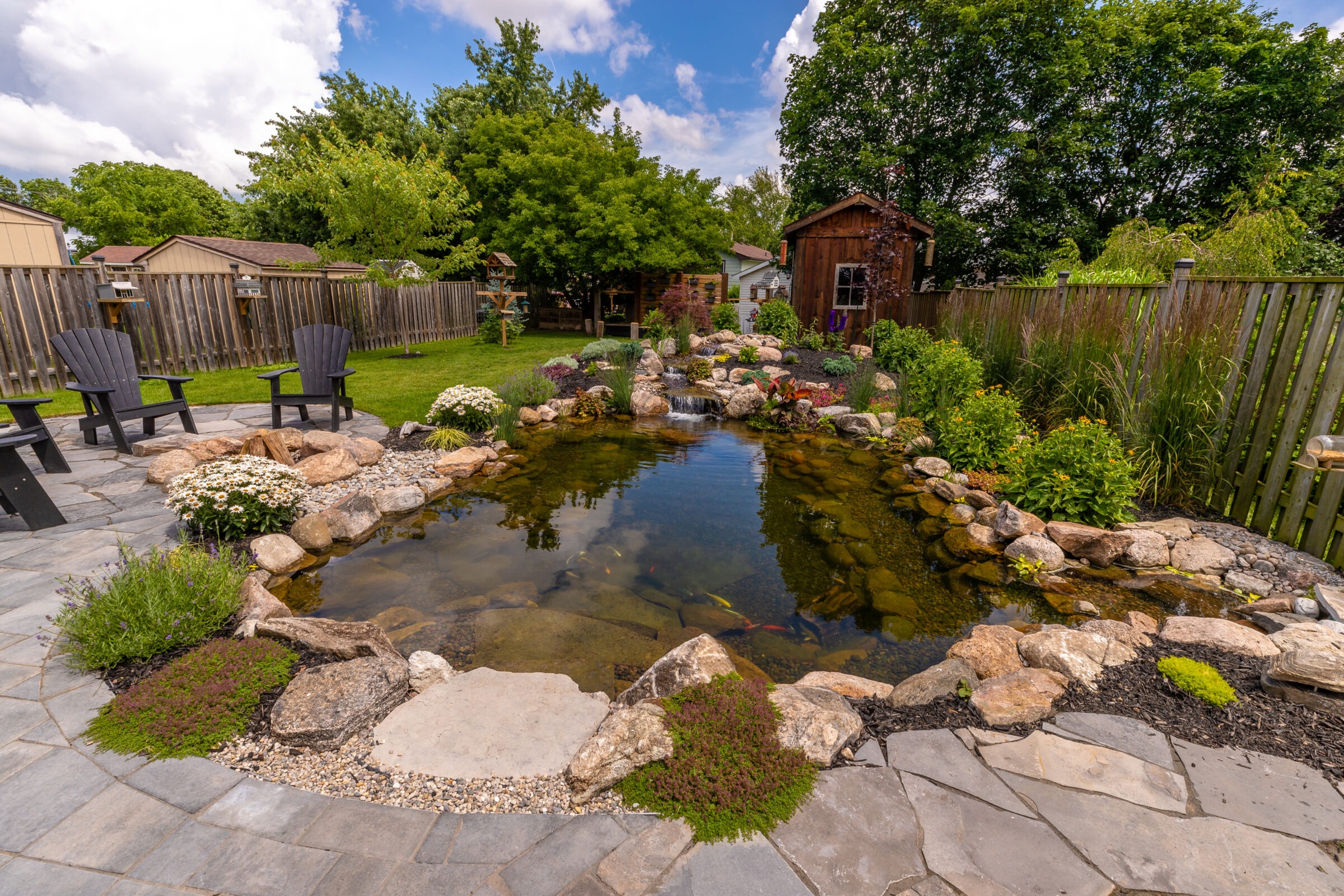 Backyard with a serene pond, surrounded by rocks and plants. Two Adirondack chairs face it, under a clear blue sky.