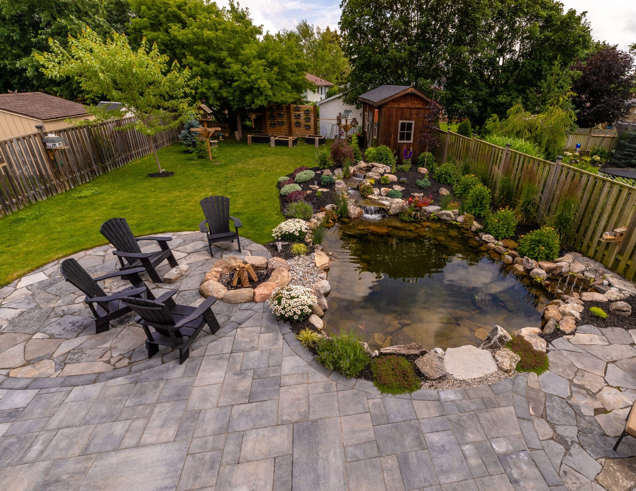 Backyard with a pond, wooden shed, rock fire pit, surrounded by greenery, stone patio, and four chairs under a partially cloudy sky.