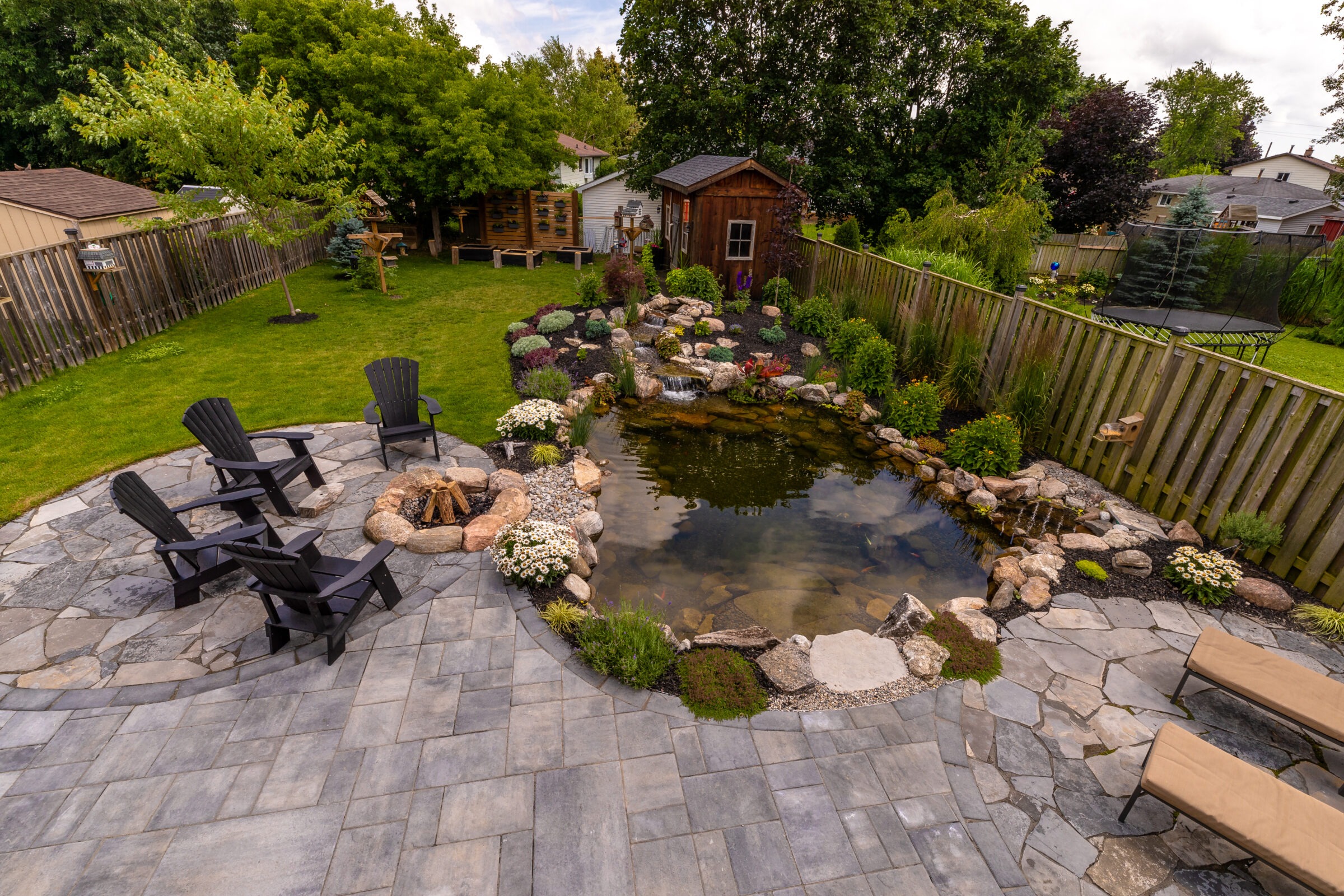 A lush garden features a pond, four chairs, fire pit, and patio, surrounded by grass, trees, and a wooden shed.