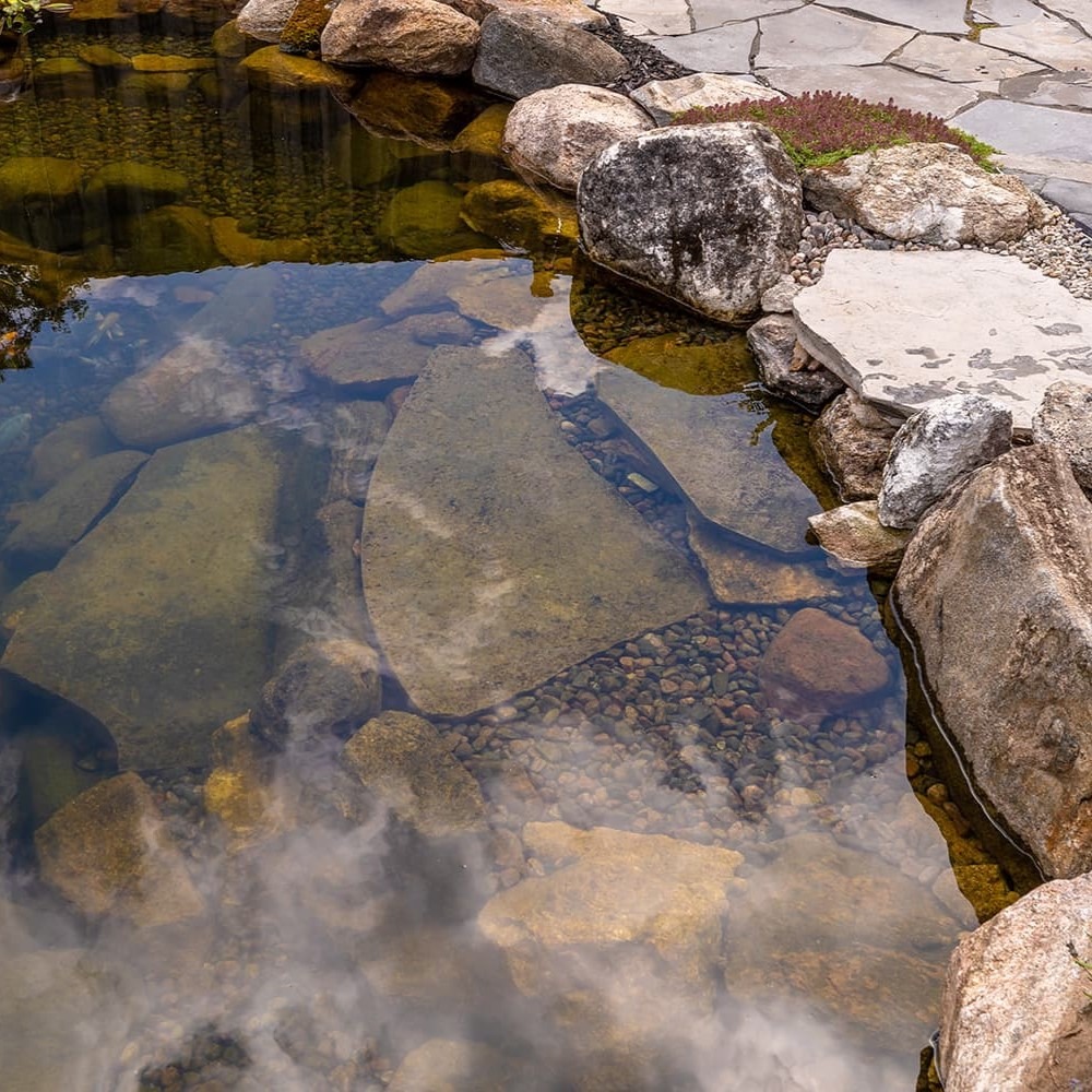 Clear pond with stones and rocks lining the edge, reflecting cloudy sky; serene natural setting with tranquil water surface.