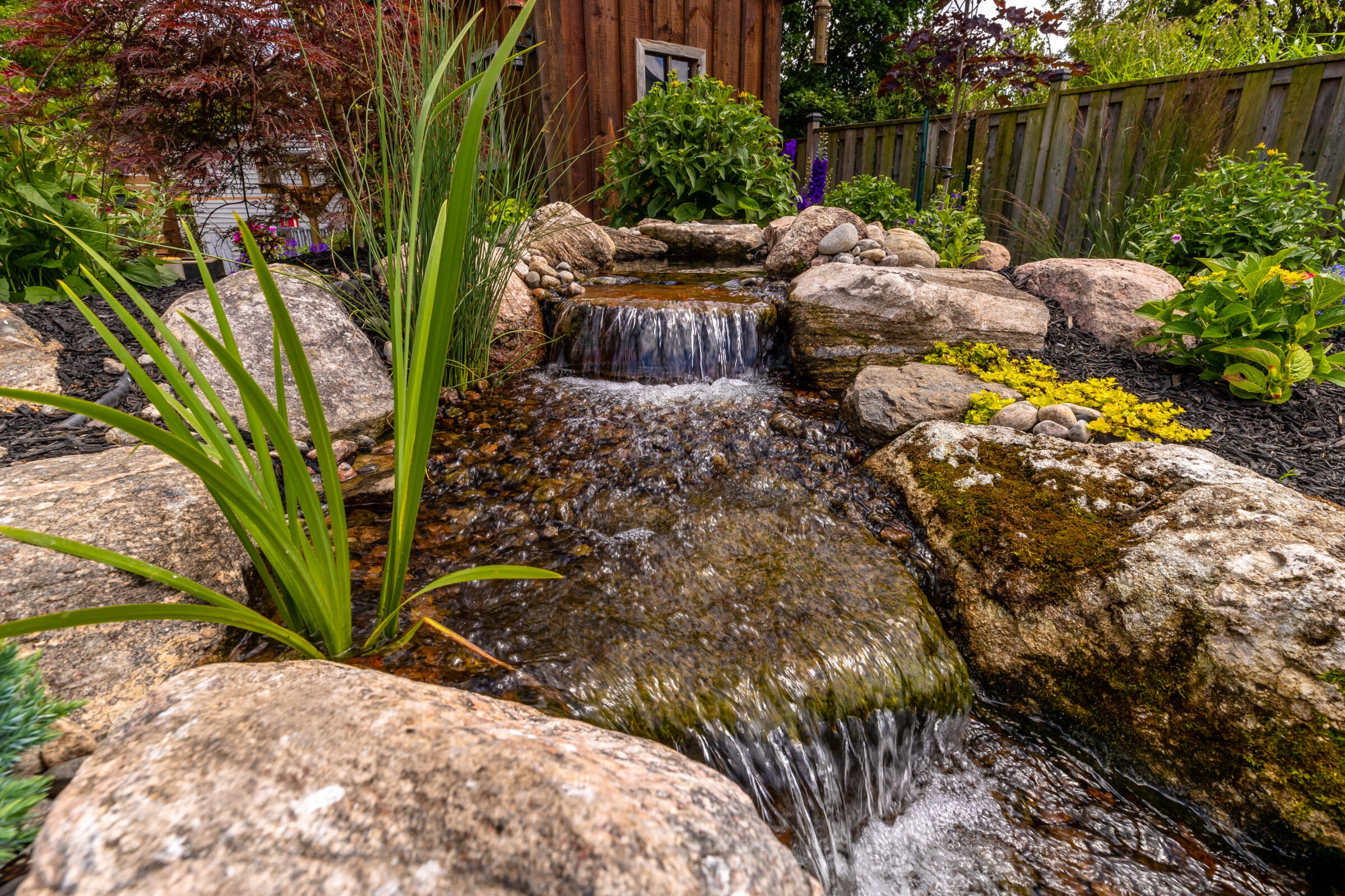 A serene garden waterfall flows over rocks, surrounded by lush greenery and a wooden fence. Plants and foliage add vibrant colors.
