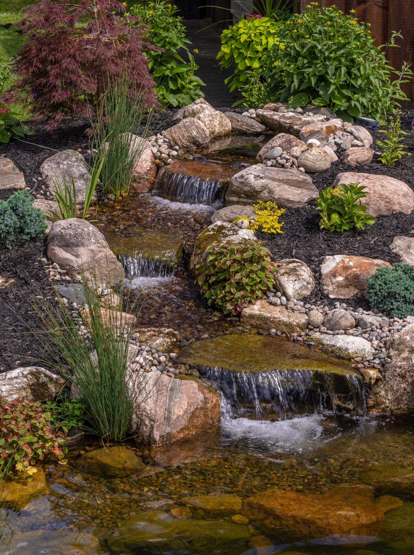 A serene garden with a small waterfall cascading over rocks, surrounded by lush greenery and colorful plants, creating a tranquil atmosphere.