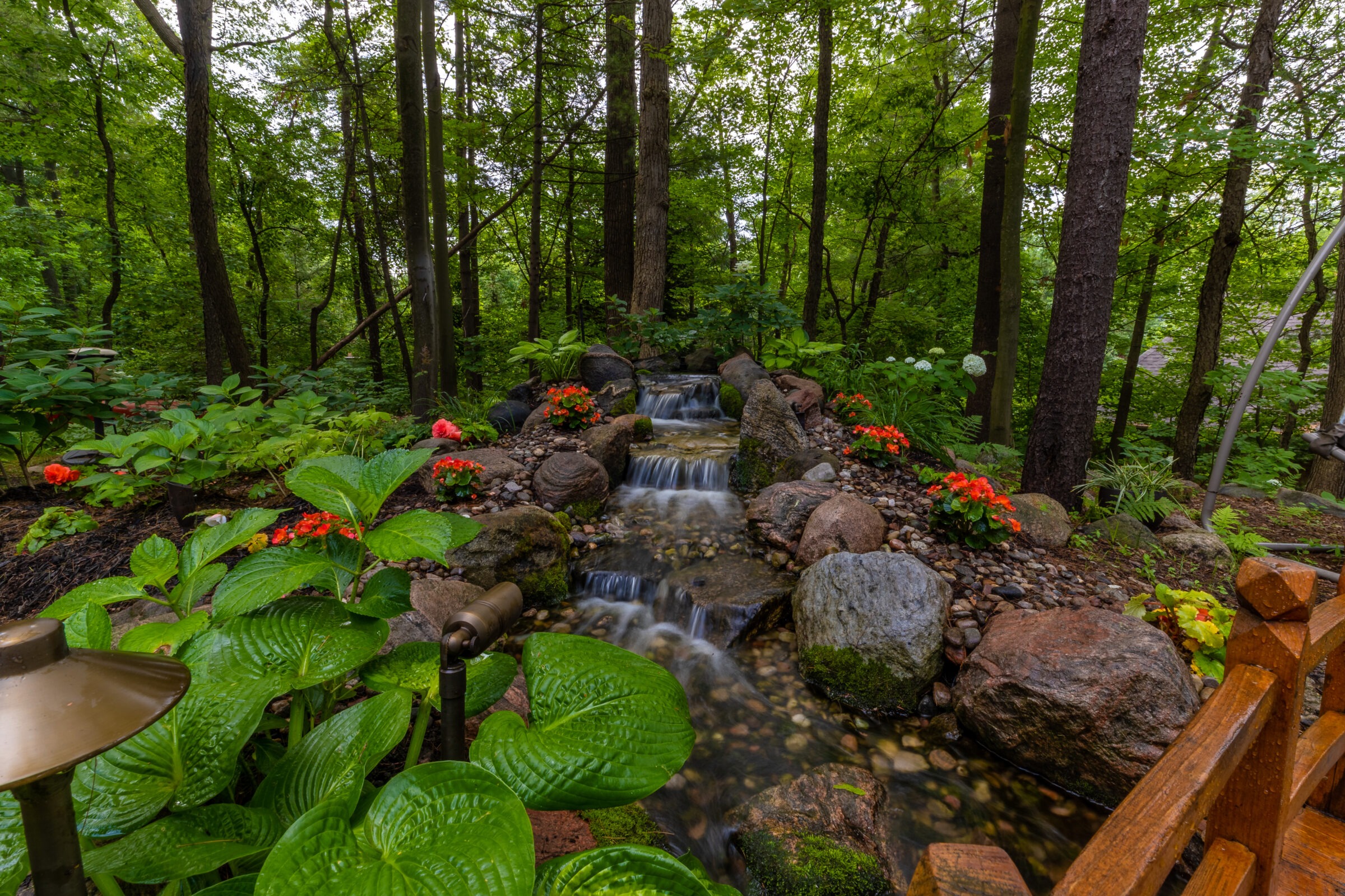 A serene forest garden features a small waterfall surrounded by lush greenery and vibrant flowers, with a wooden structure partially visible.
