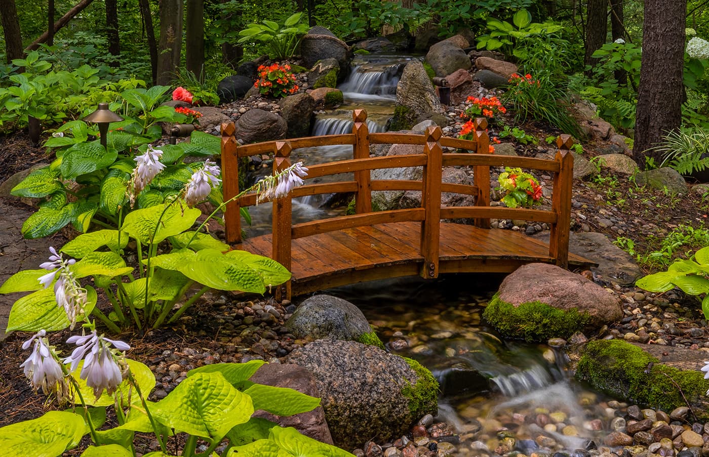 A serene garden scene with a small wooden bridge over a stream, surrounded by vibrant flowers, lush greenery, and rocks.