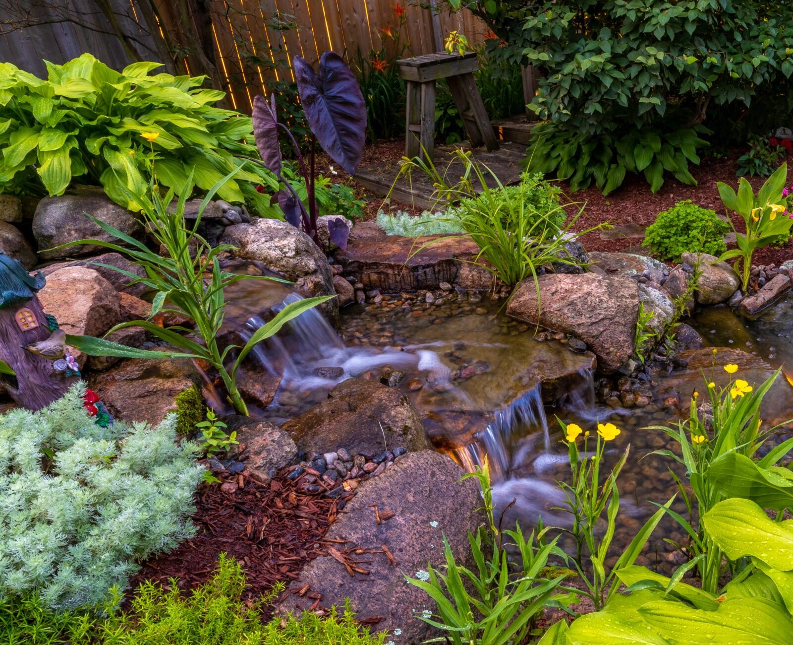 A serene garden scene featuring a small waterfall, vibrant greenery, colorful flowers, pond with lily pads, and decorative rock arrangements.