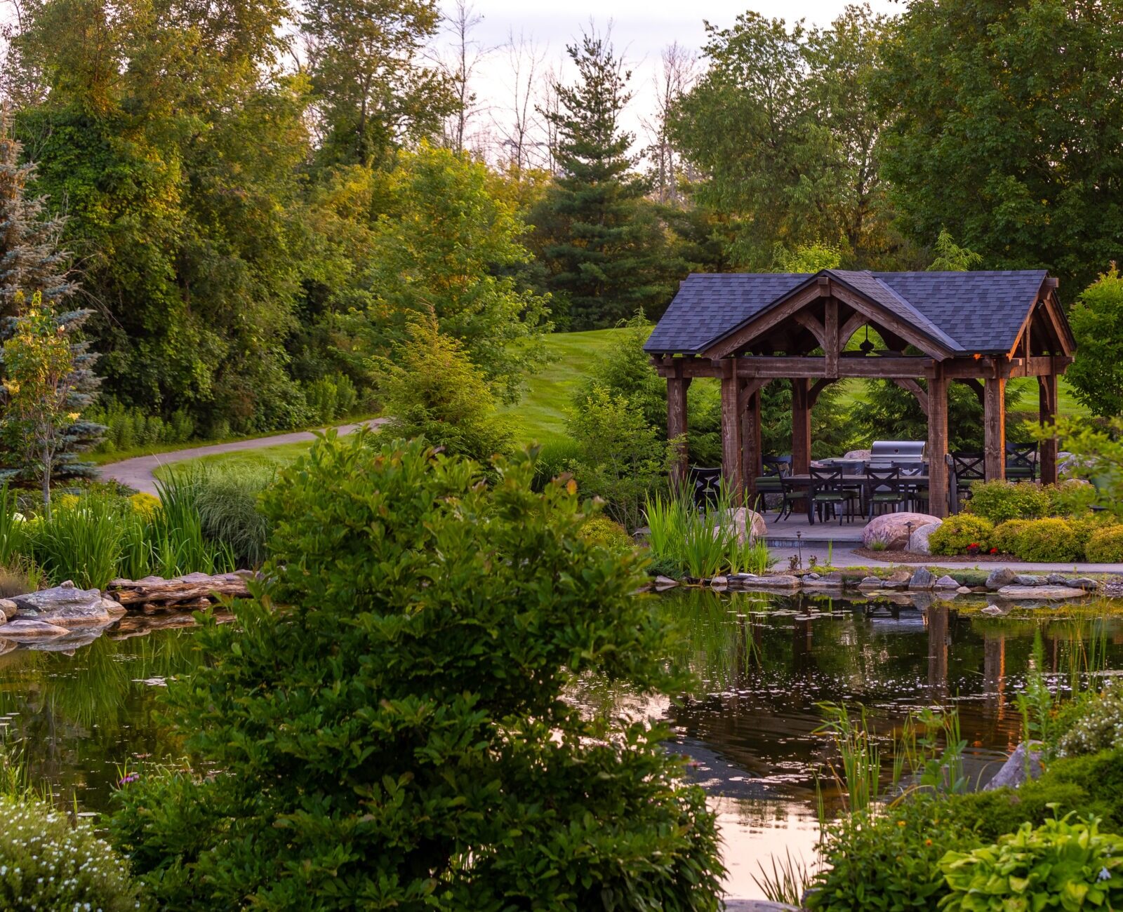 A serene garden scene with a wooden gazebo next to a pond, surrounded by lush greenery and a winding path.