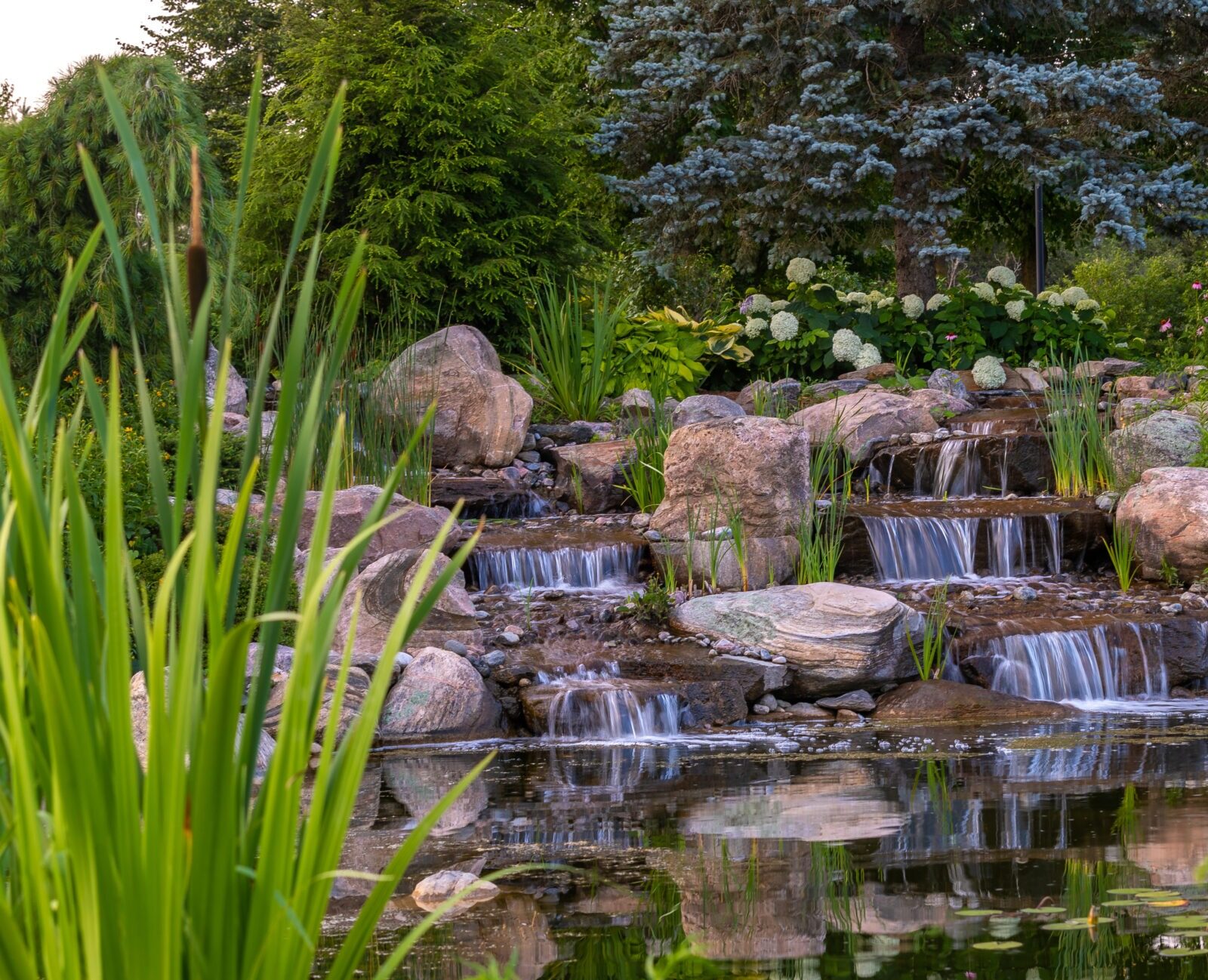 A serene garden scene features a cascading waterfall with rocks, surrounded by lush greenery and colorful flowers reflecting in a tranquil pond.