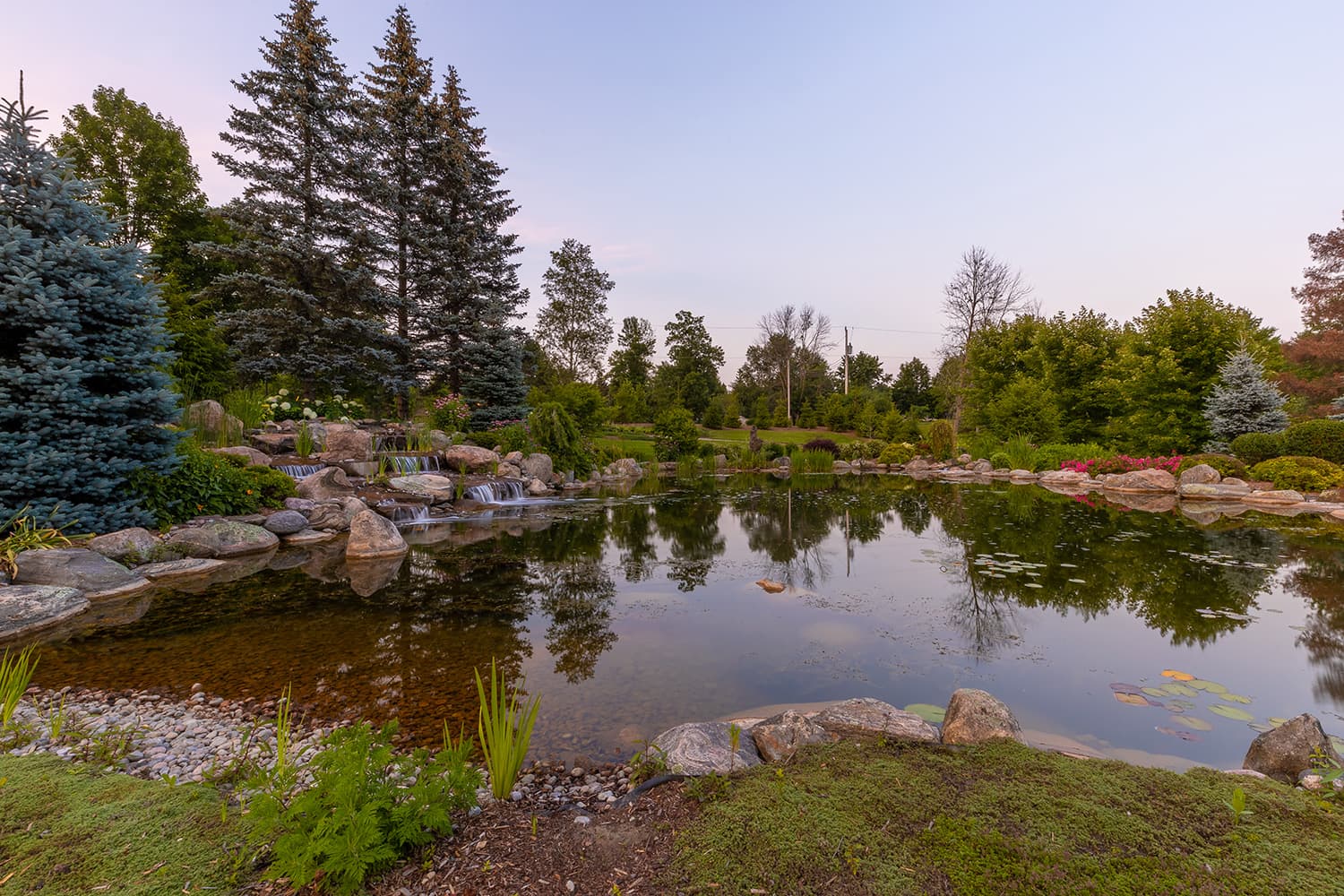 A serene pond with cascading waterfalls, surrounded by lush greenery and trees, reflecting a clear blue sky during a tranquil day.