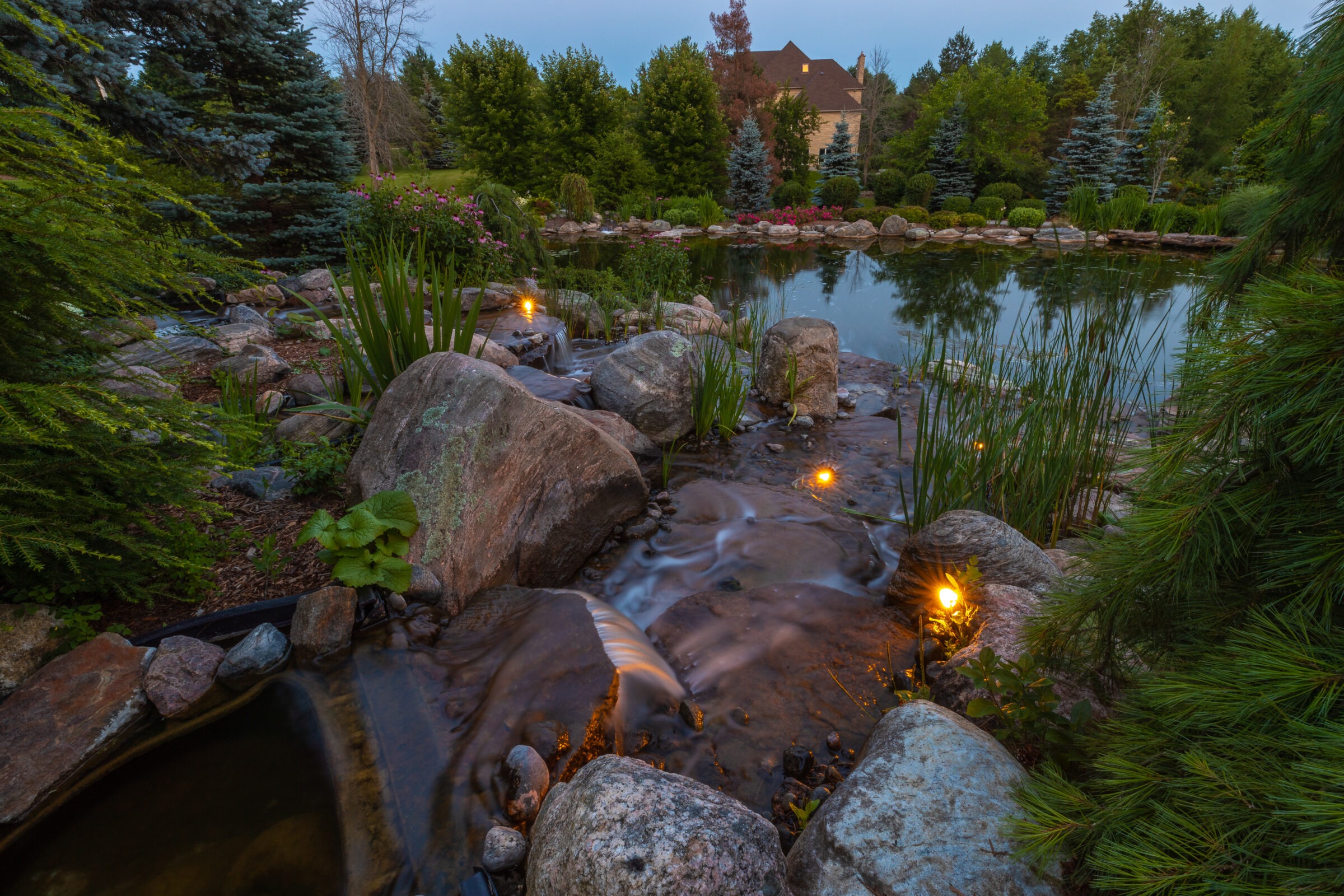 Tranquil garden setting with a small waterfall, illuminated by lights, leading to a pond surrounded by lush greenery and trees in the background.