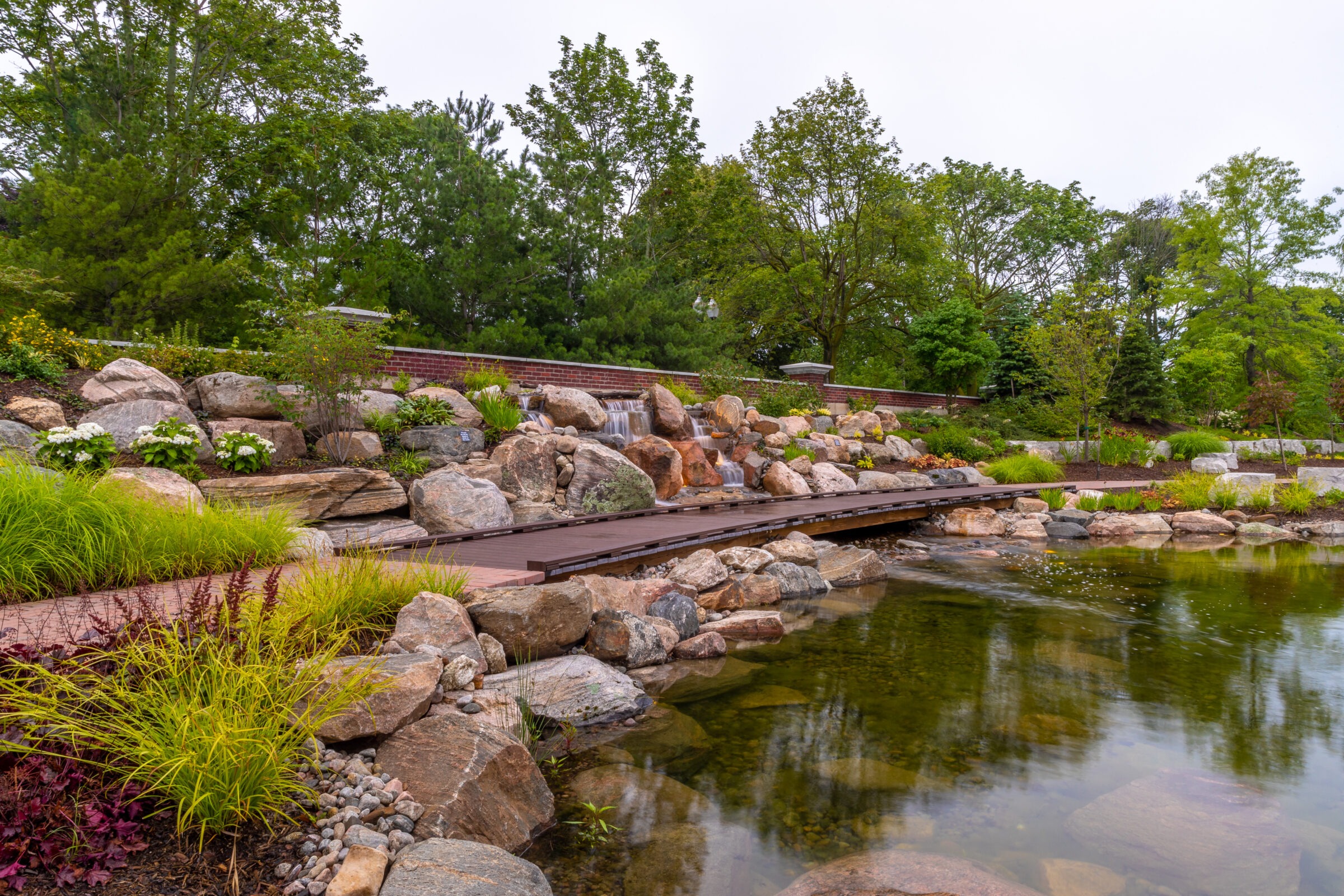 Tranquil garden with cascading waterfall, rocks, lush greenery, and a small wooden bridge over a clear pond, surrounded by trees.