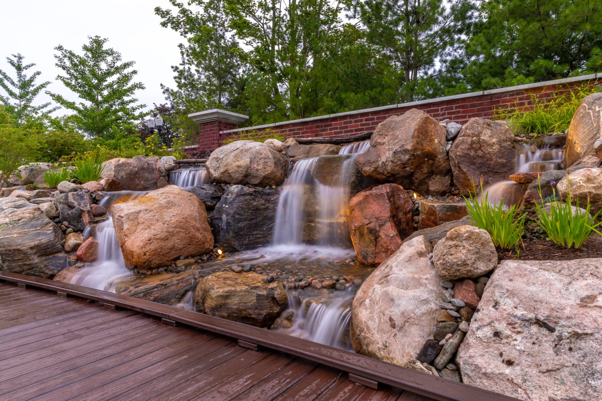 A serene waterfall flows over rocks into a small stream, surrounded by lush greenery and a brick wall in a garden setting.