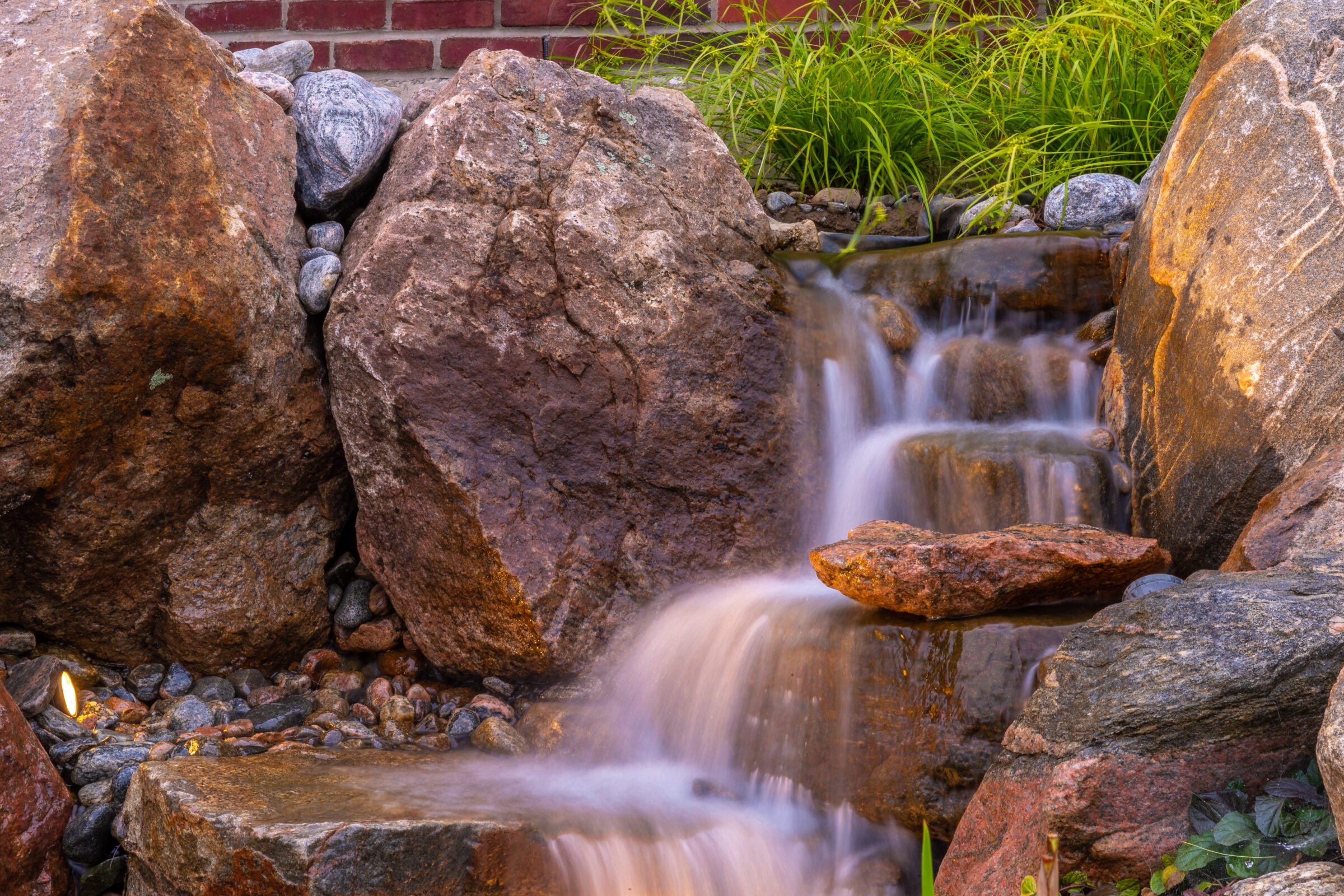 Water cascades gently over smooth rocks surrounded by lush green plants, creating a tranquil and serene natural atmosphere.