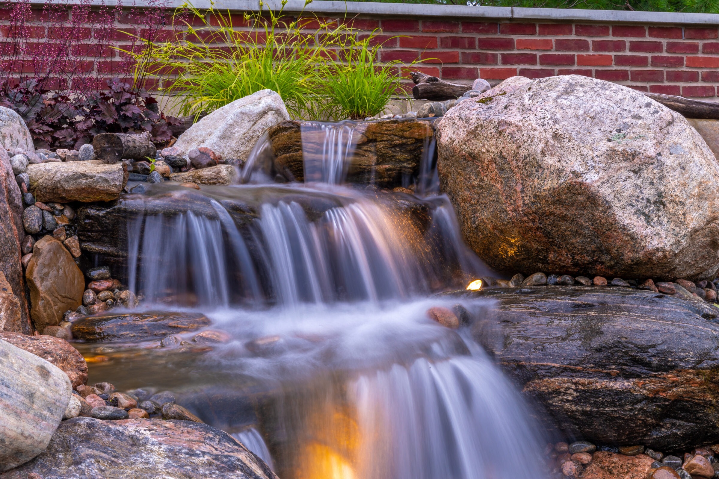 Water cascades gently over rocks, surrounded by lush greenery and a red brick wall in a serene garden setting.