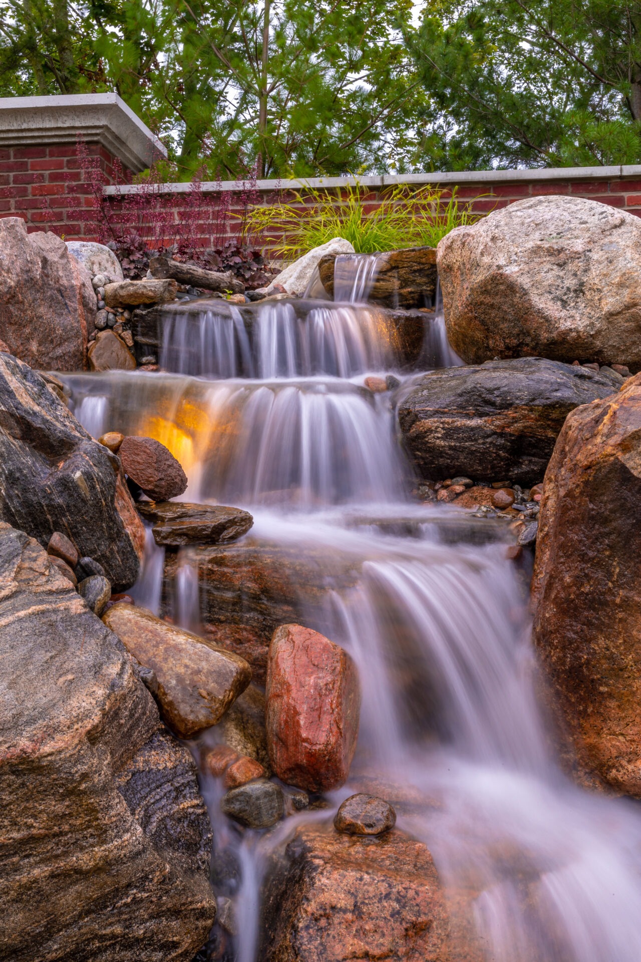 A serene waterfall cascades over smooth rocks, surrounded by lush greenery. A brick wall with foliage adds to the tranquil garden setting.