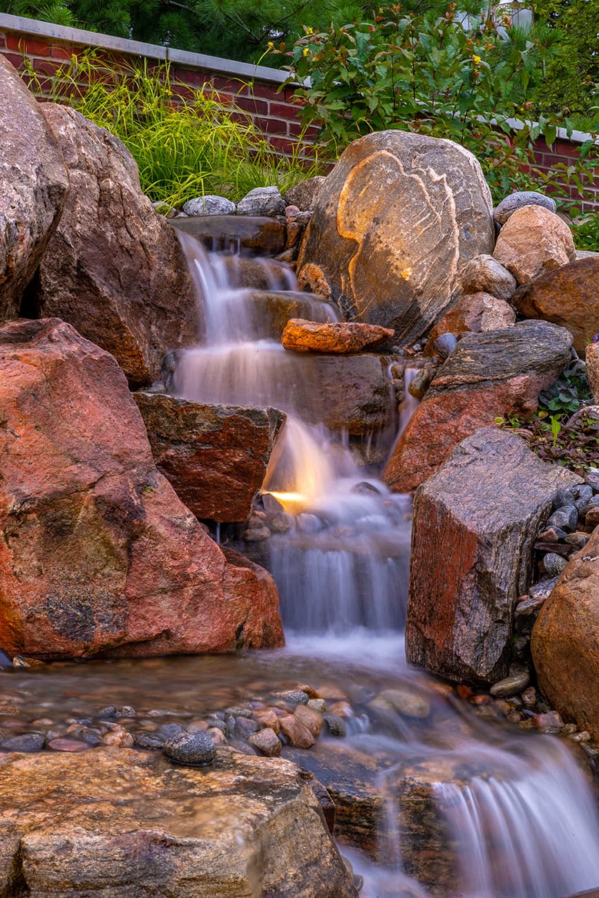 A small waterfall flows between large, textured rocks, surrounded by green plants. Soft lighting highlights the natural scene, creating a tranquil atmosphere.