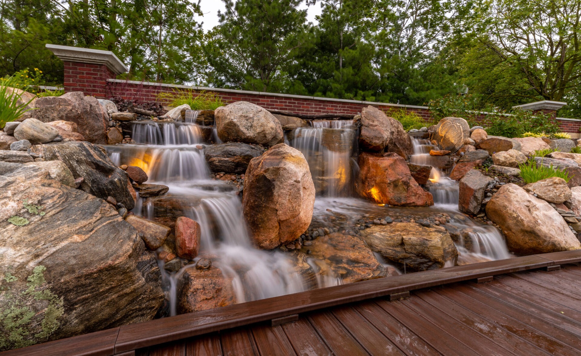 A serene, landscaped waterfall flows over rocks, surrounded by greenery, with a wooden deck in the foreground and a brick wall backdrop.