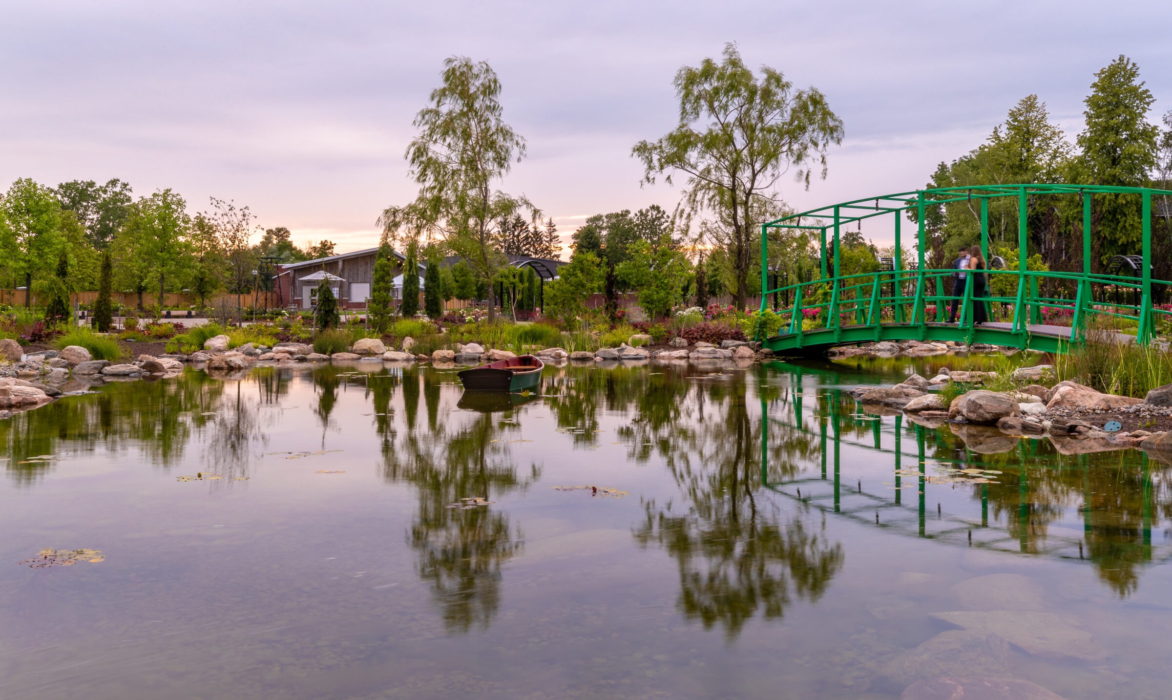 Peaceful park scene with a green bridge, lush trees, pond reflections, and two people on bridge. Calm water with scattered stones.