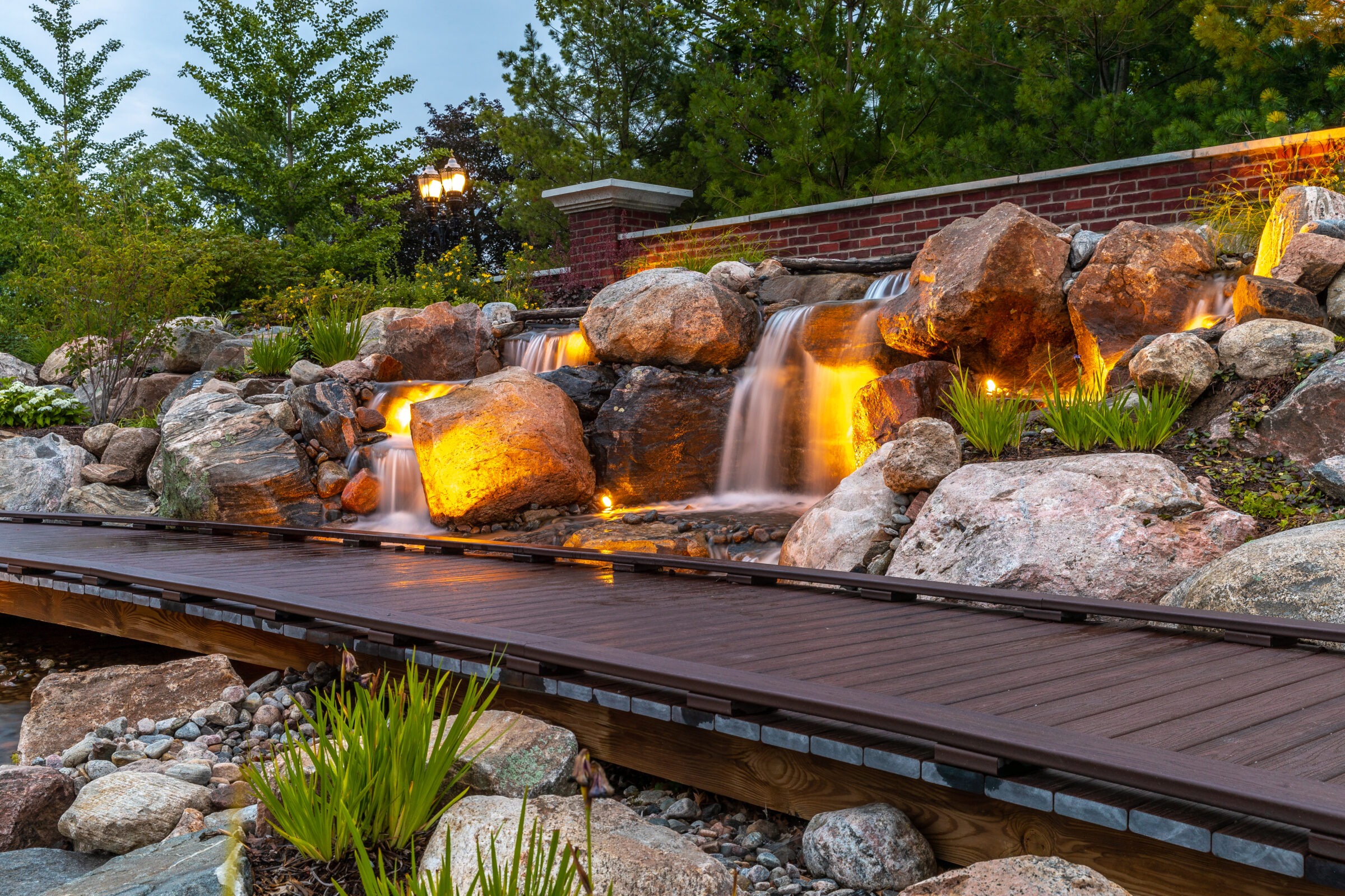 Illuminated stone waterfall flows beside a wooden walkway, surrounded by lush greenery and a brick wall, creating a serene, natural landscape setting.