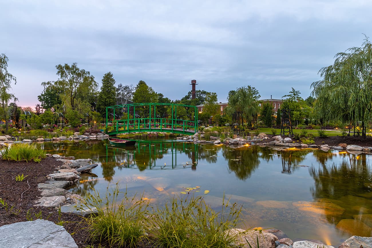 A serene park features a vibrant green bridge over a reflective pond, surrounded by lush greenery and a distant smokestack.