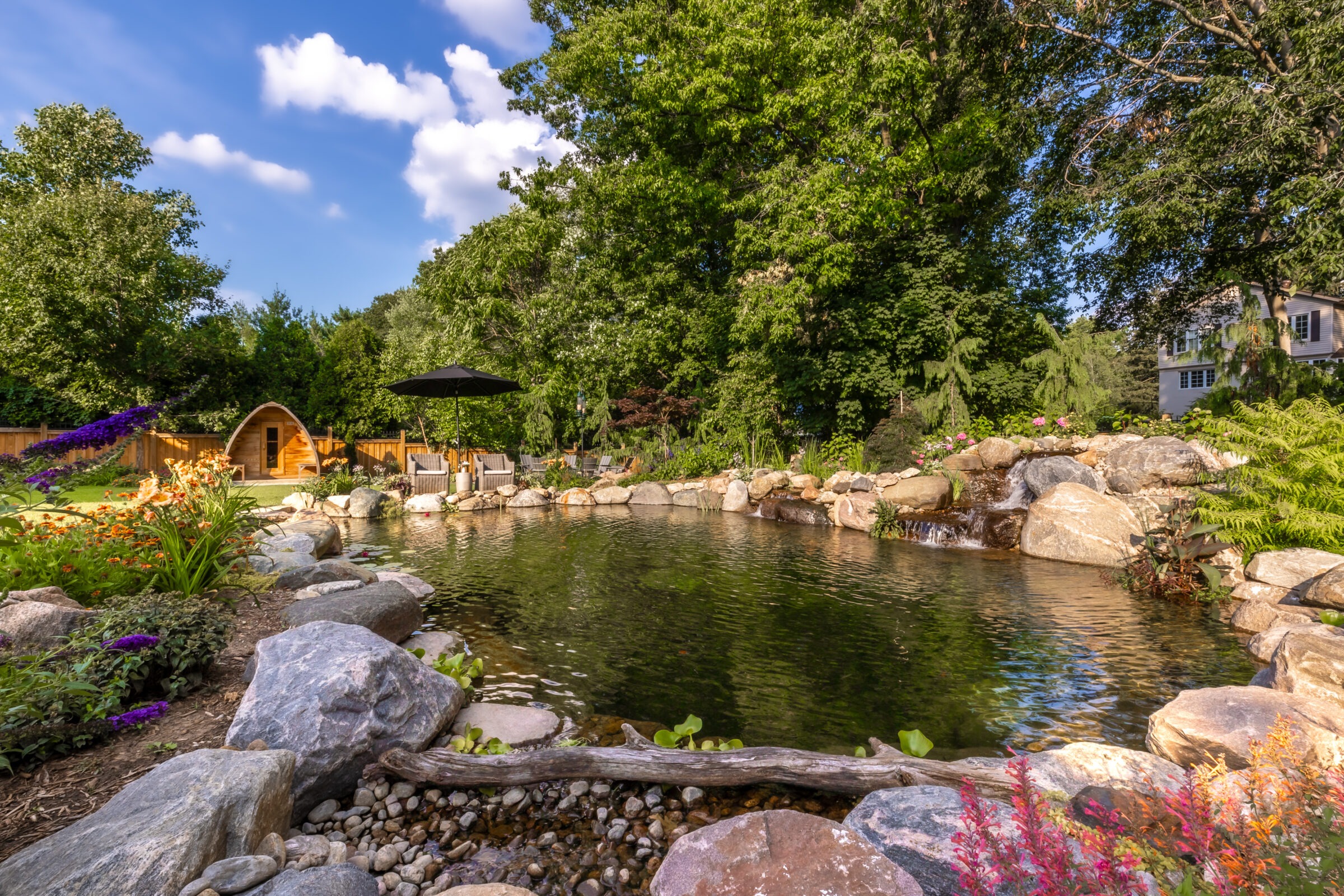 A serene garden features a natural pond with colorful flowers, surrounded by trees. Small wooden structure and seating area under a black umbrella.