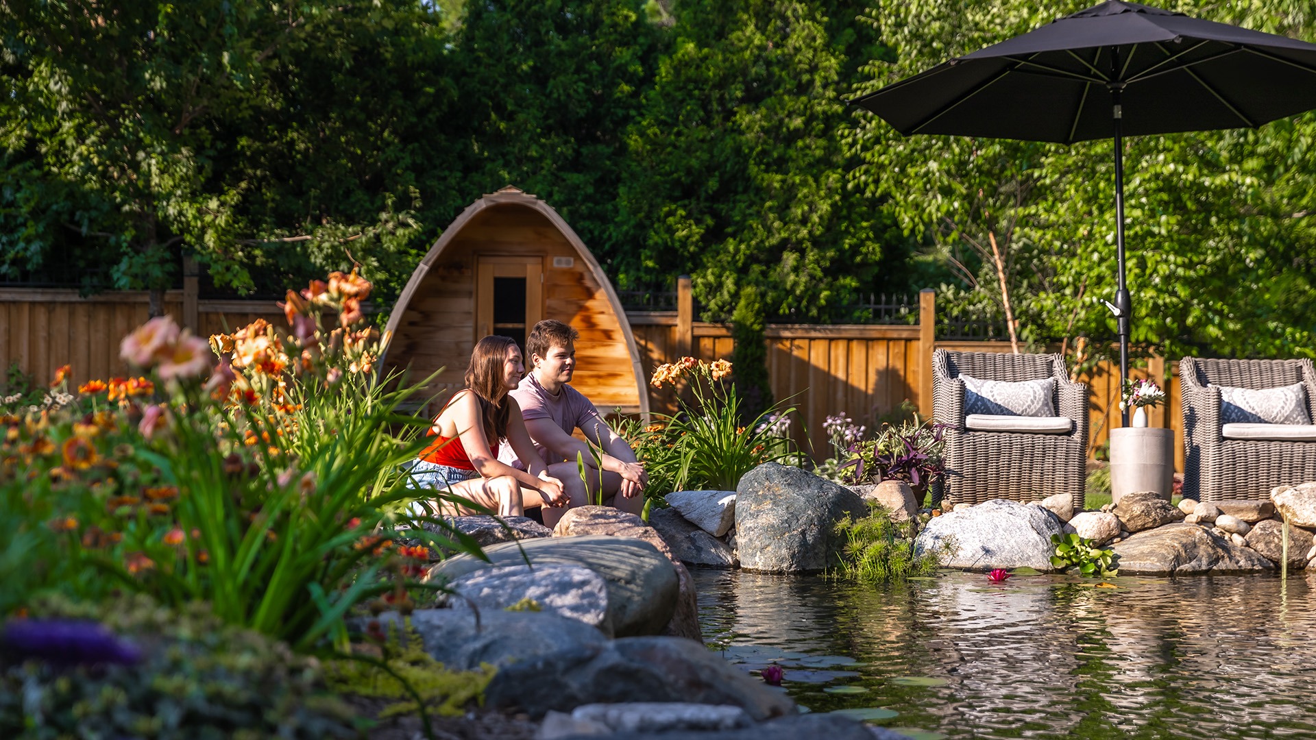 Two people sit by a tranquil garden pond, surrounded by flowers, rocks, and greenery, next to a small wooden cabin and patio furniture.