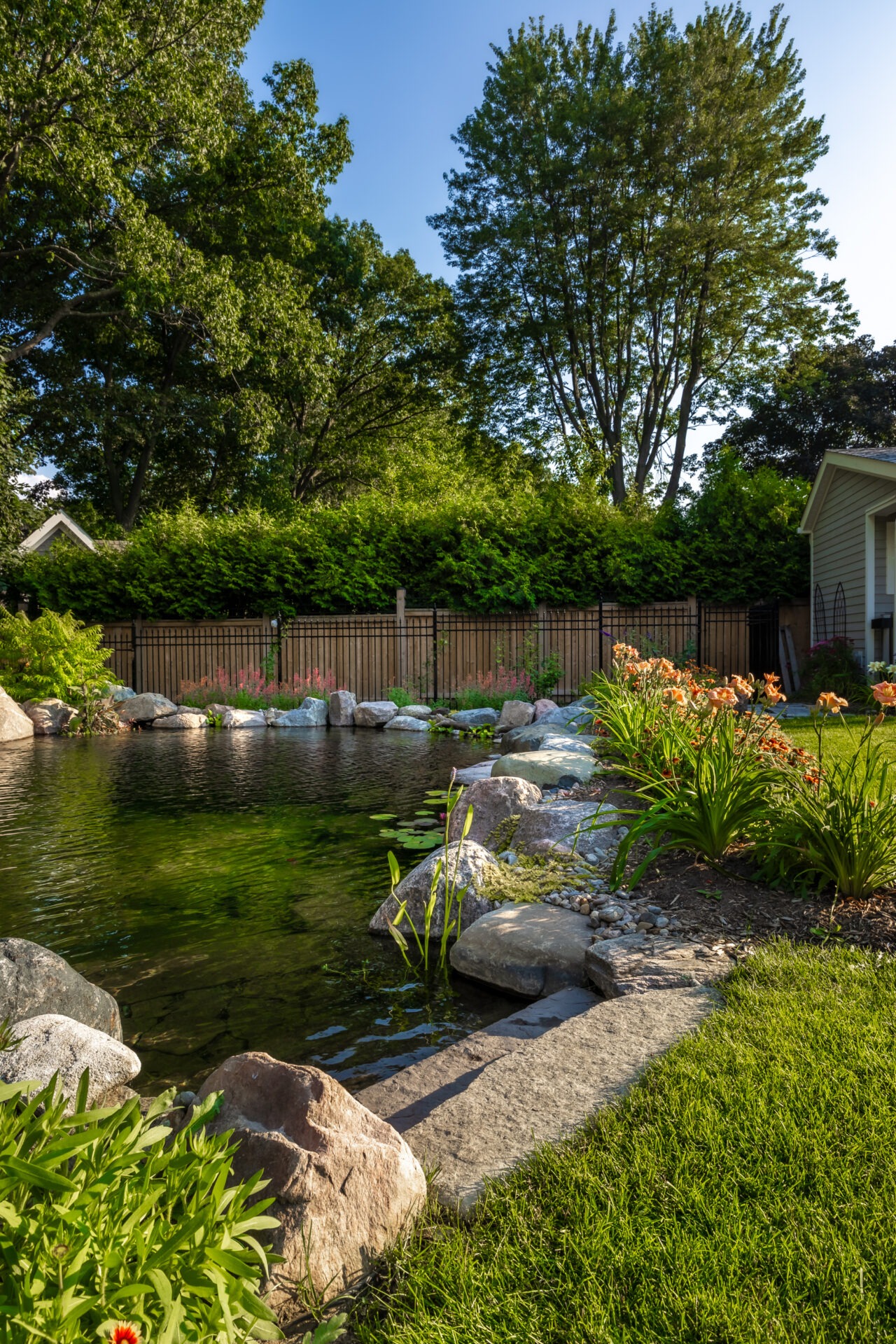 A serene garden scene with a rock-lined pond, colorful flowers, vibrant greenery, and tall trees under a clear blue sky.