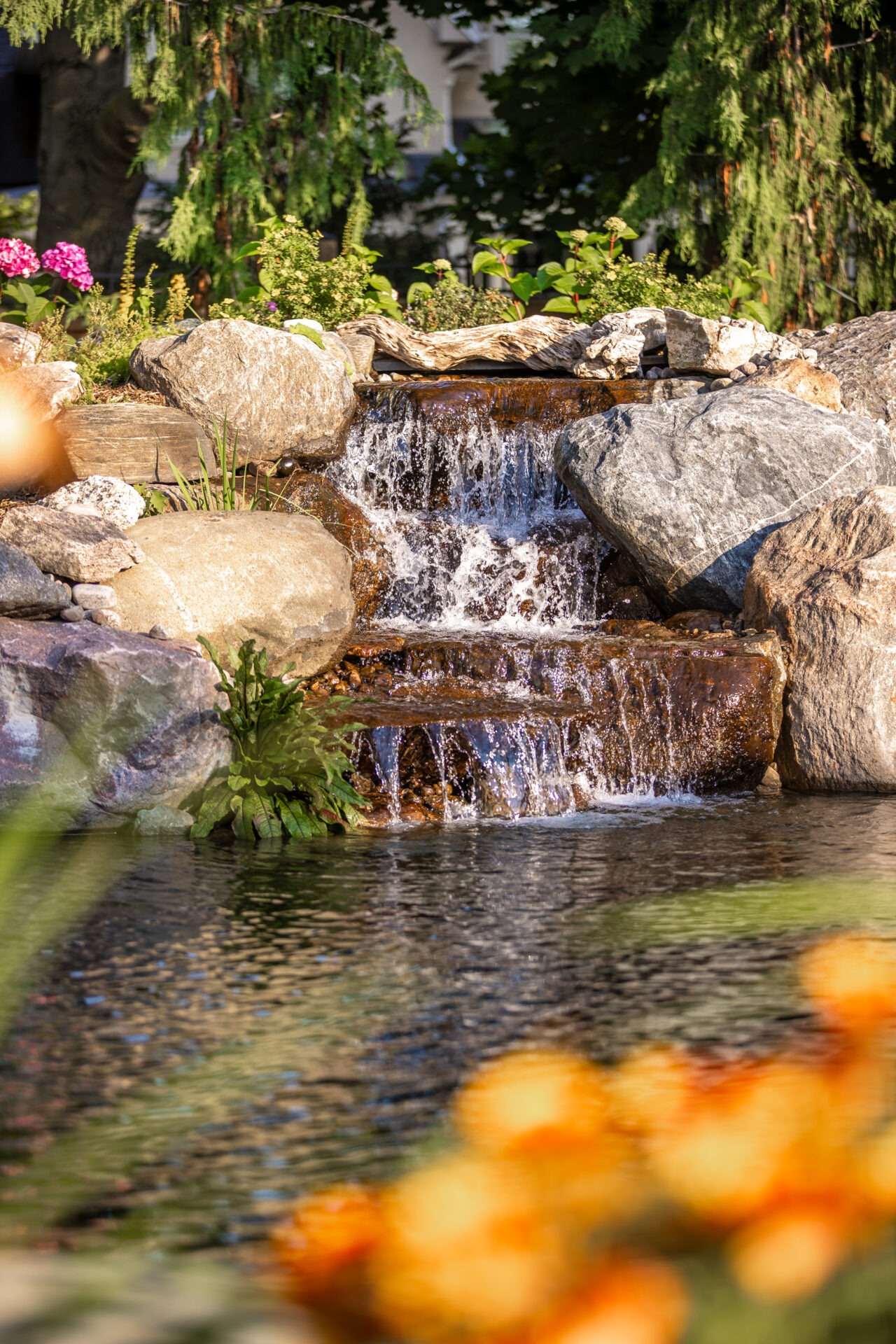 Tranquil garden waterfall surrounded by rocks and greenery, with colorful flowers in the foreground, creating a peaceful nature scene.