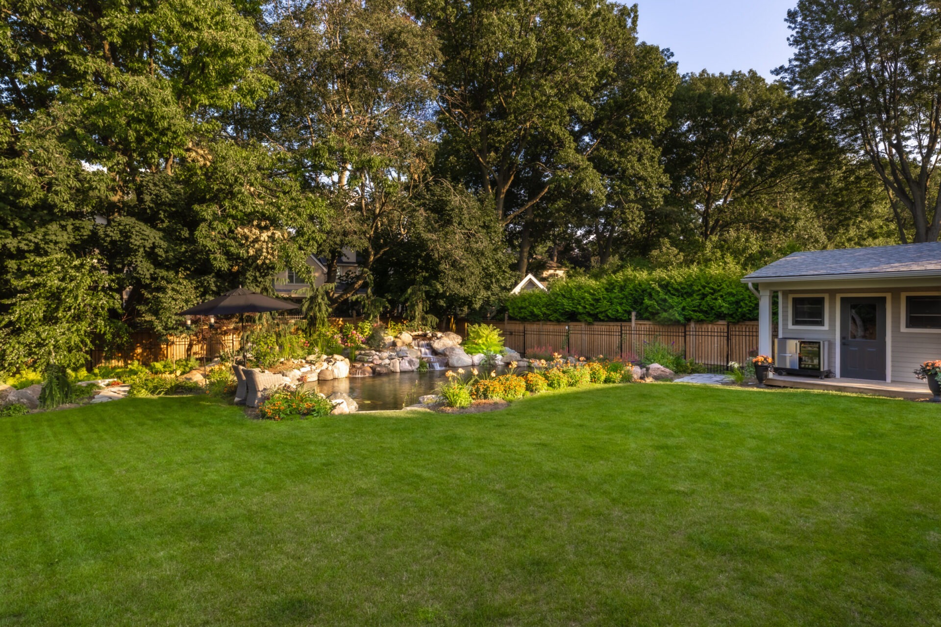 A serene backyard with green lawn, vibrant flower garden, small pond with rocks, surrounded by tall trees, adjacent to a cozy shed.