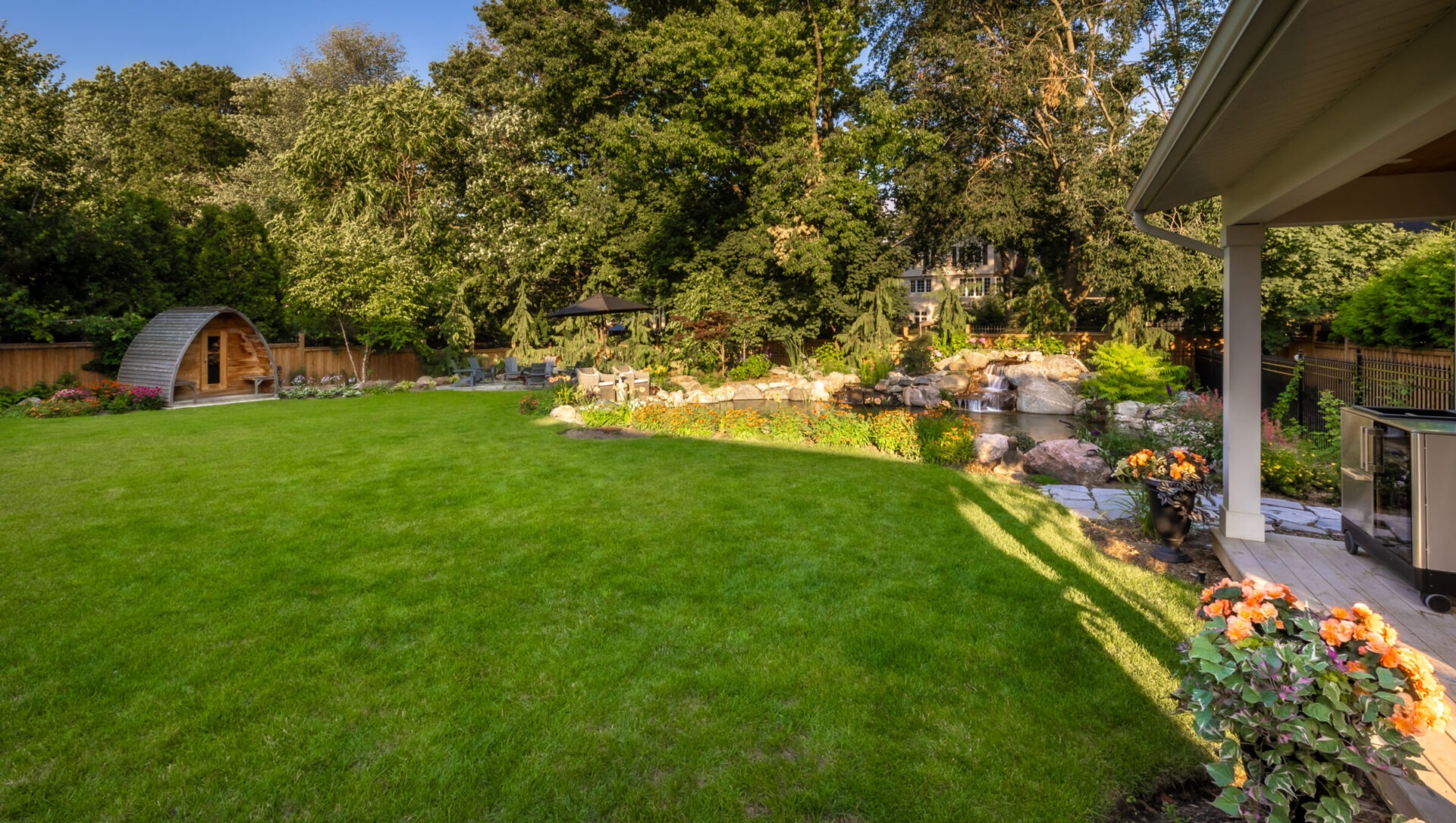 Lush backyard garden with vibrant flowers, a pond, and a wooden sauna hut. Trees and shrubs provide privacy, creating a serene atmosphere.