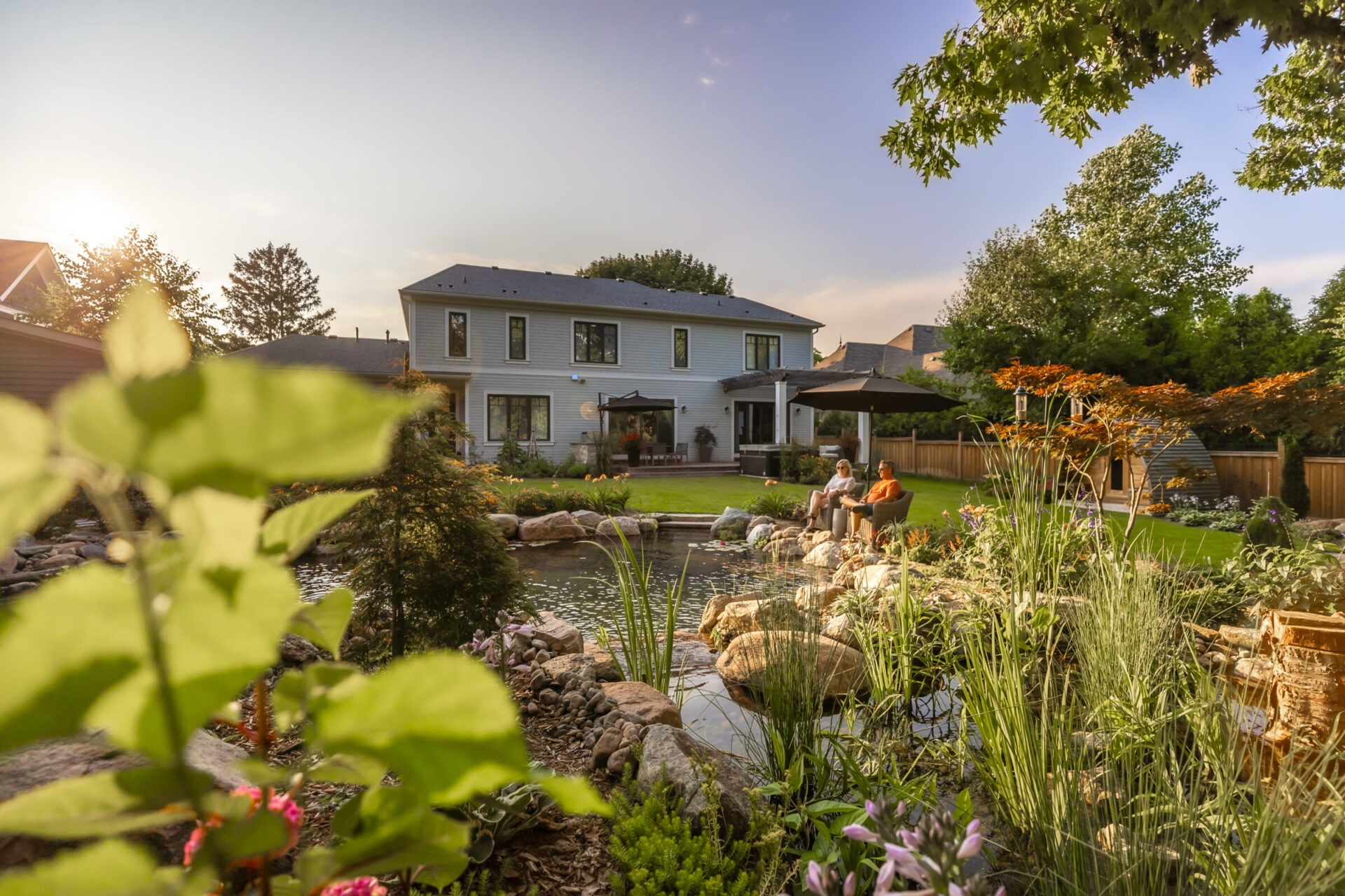 Two people sit by a garden pond in front of a house, surrounded by plants and trees under a clear sky.