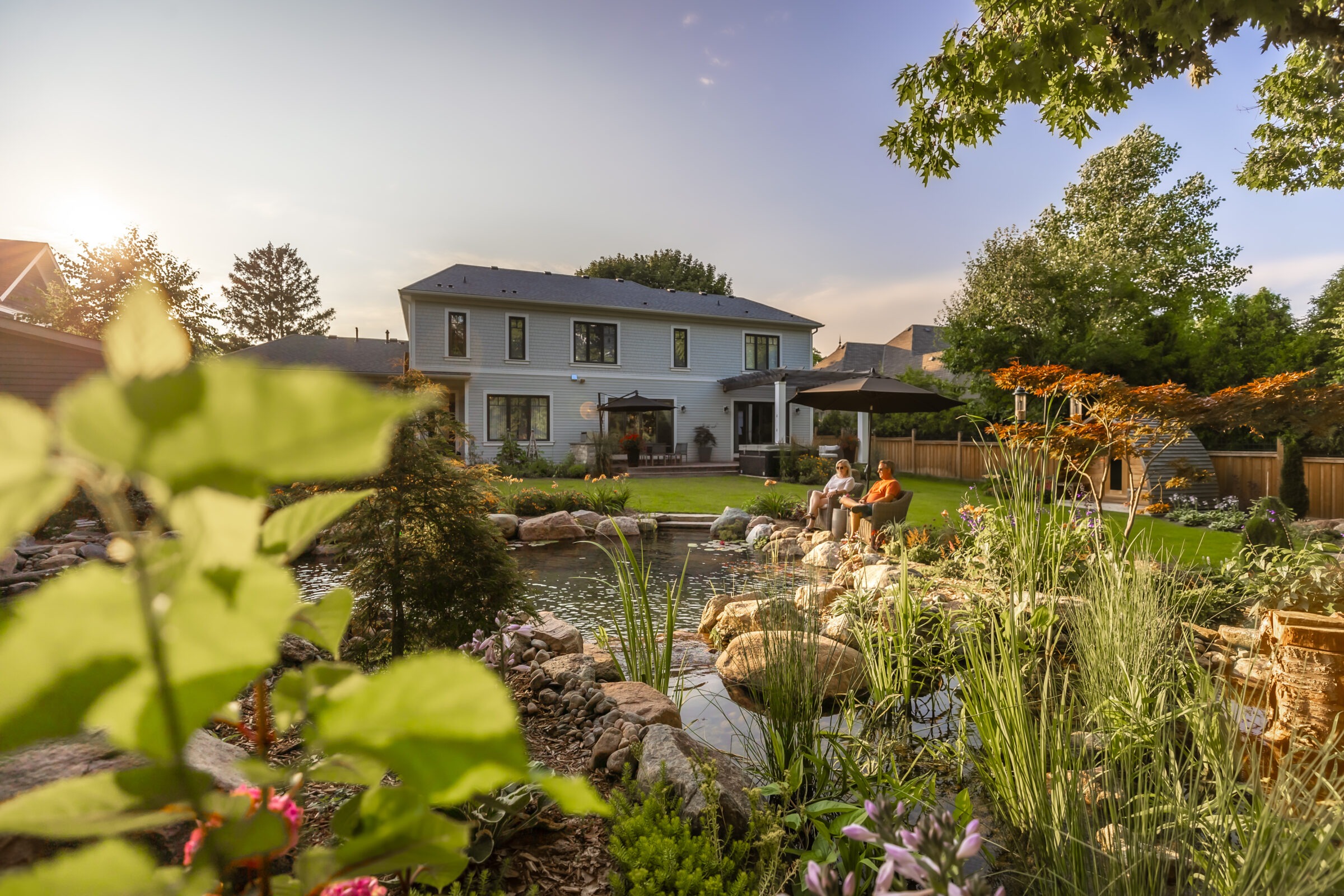 Two people sit by a garden pond in front of a house, surrounded by plants and trees under a clear sky.