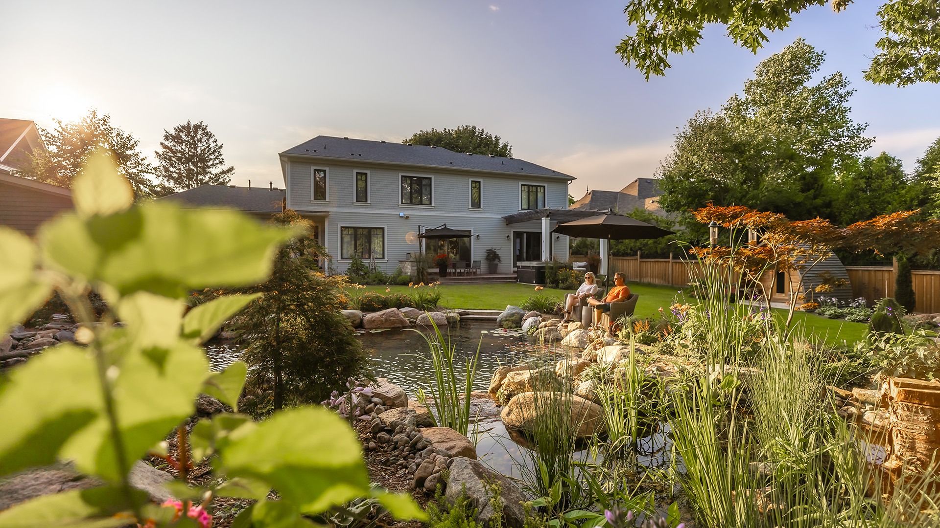 A serene backyard scene with two people relaxing by a pond. Lush greenery and a modern house create a peaceful atmosphere.