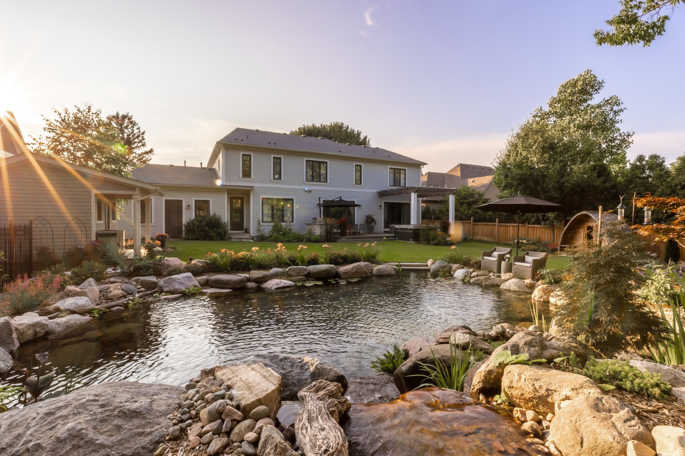 Large house with a well-maintained backyard pond, surrounded by rocks and plants, under a clear sky with the sun setting.