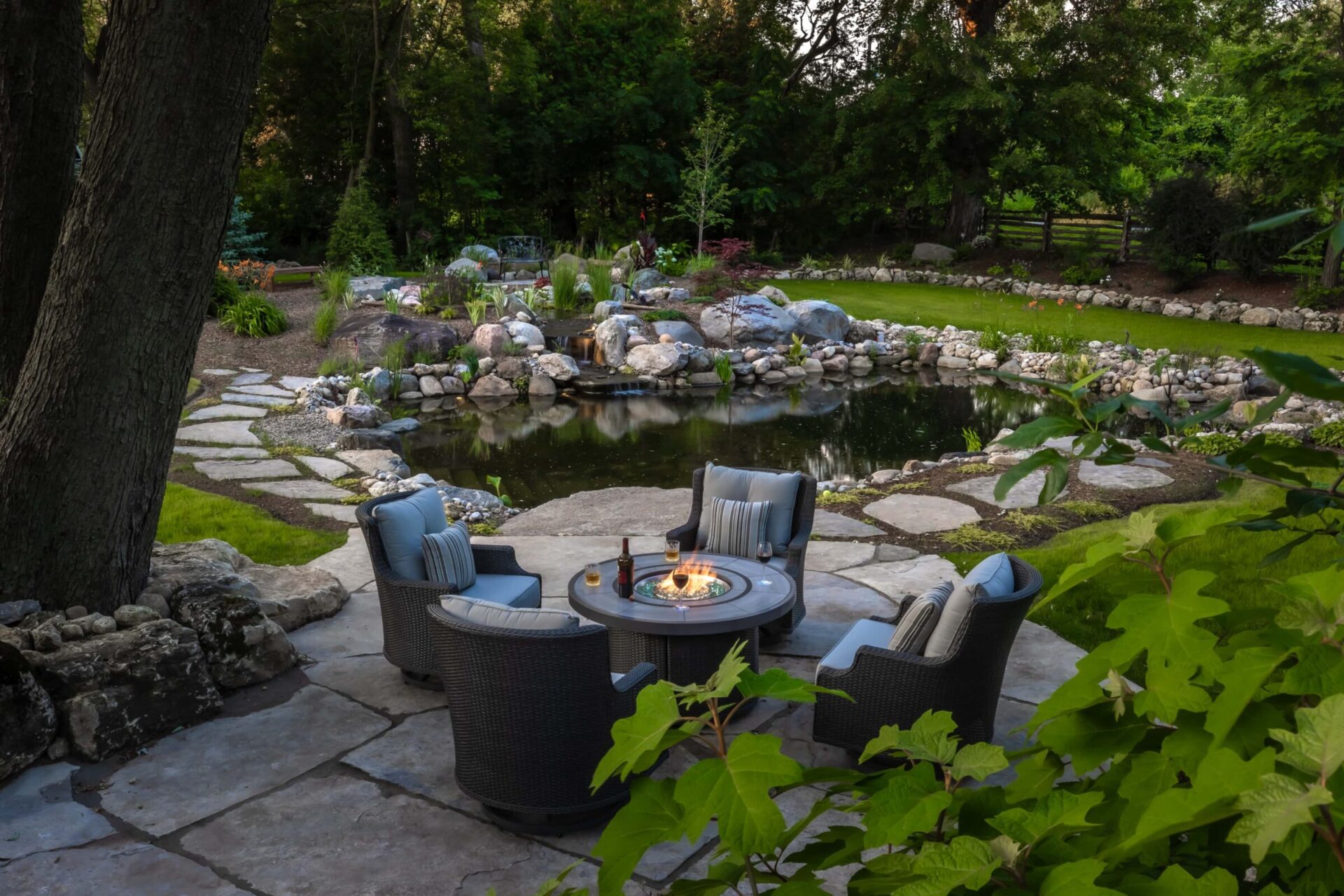 Tranquil garden scene featuring a circular fire pit, four cushioned chairs, and a picturesque pond surrounded by lush greenery and stone pathways.