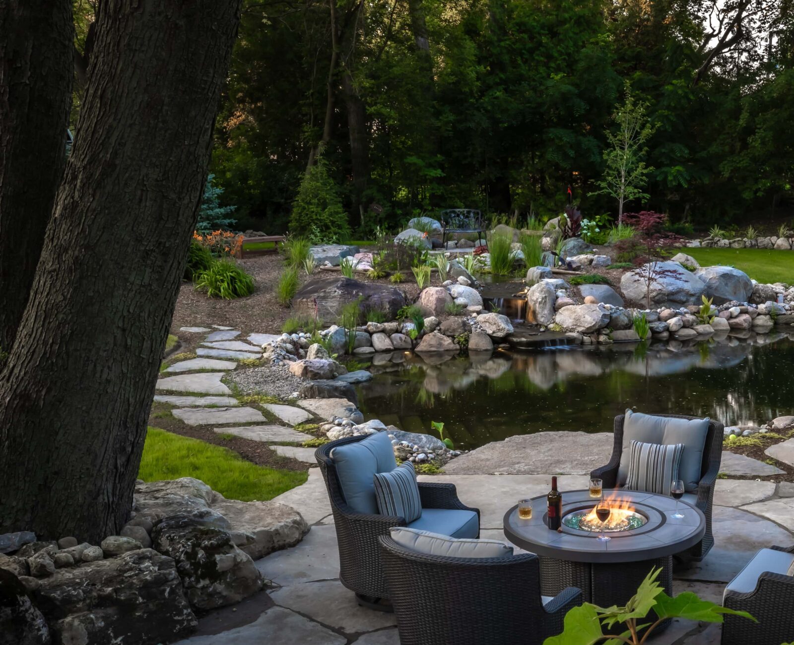 Tranquil garden scene featuring a circular fire pit, four cushioned chairs, and a picturesque pond surrounded by lush greenery and stone pathways.