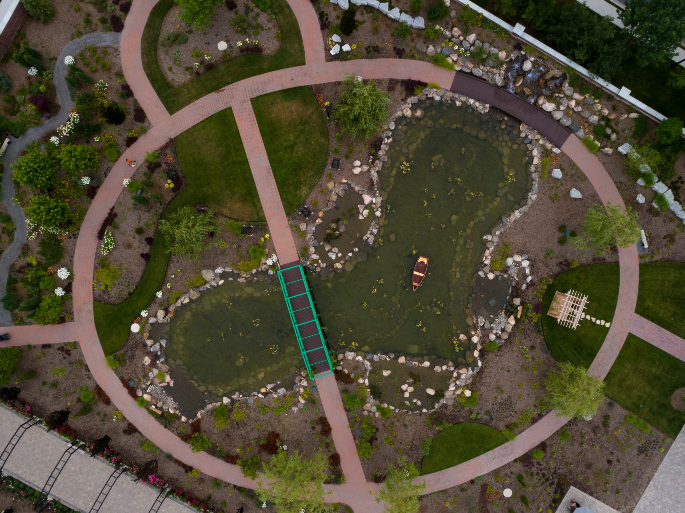 Aerial view of a landscaped garden with circular pathways, a pond, small boat, and wooden bridge, surrounded by various plants and trees.