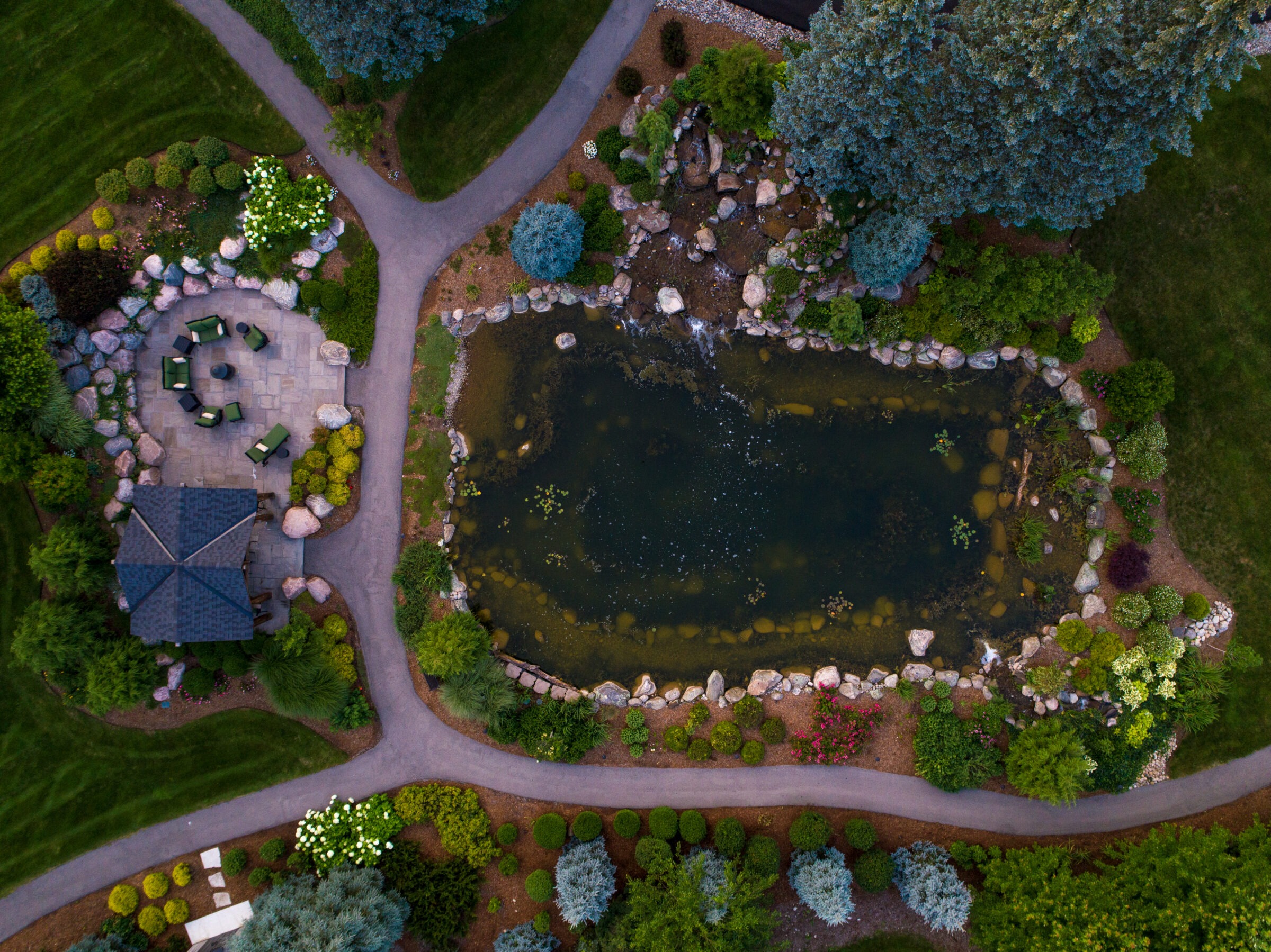Aerial view of a landscaped garden with pathways, a pond, and seating area. Lush greenery and rocks adorn the peaceful setting.