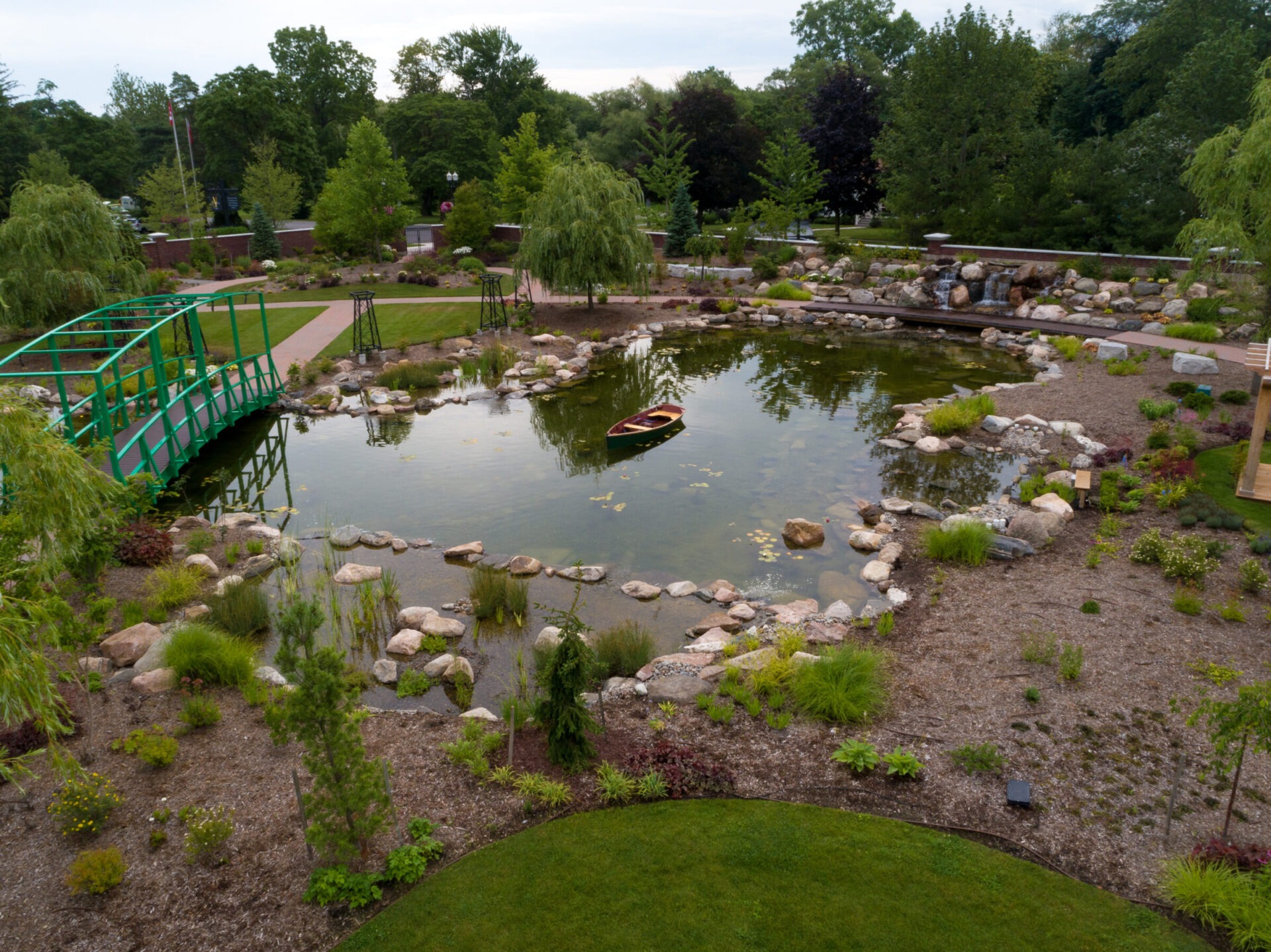A serene garden scene with a pond, green bridge, small boat, and lush plants, surrounded by trees and pathways.