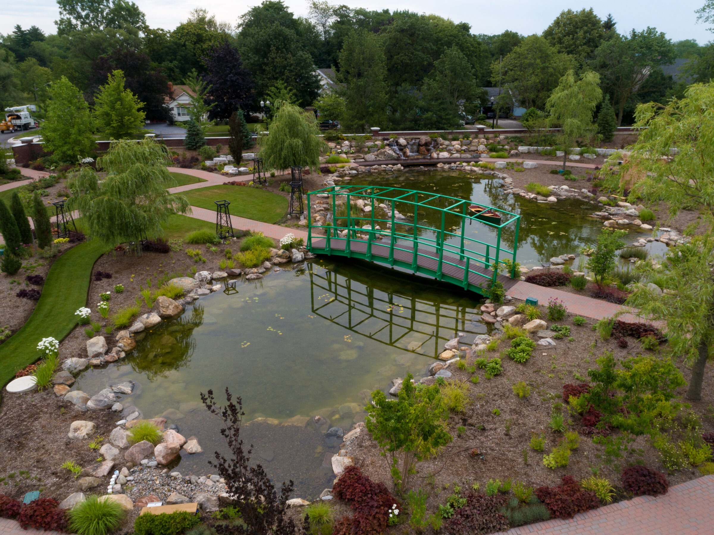 A lush garden features a green bridge over a pond, surrounded by pathways, trees, and carefully arranged rocks under a cloudy sky.
