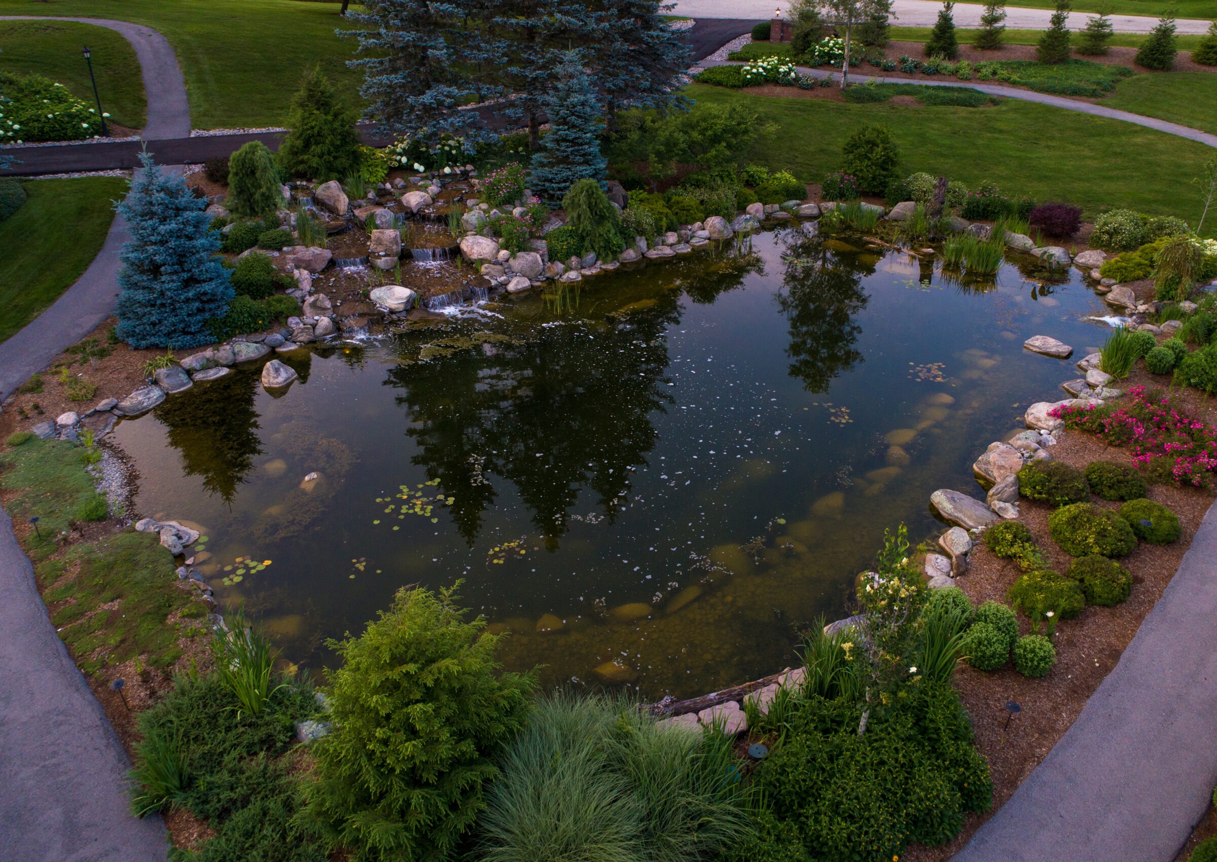 A serene garden pond with graceful pathways, surrounded by lush greenery and colorful flowers, reflecting surrounding trees in tranquil water.